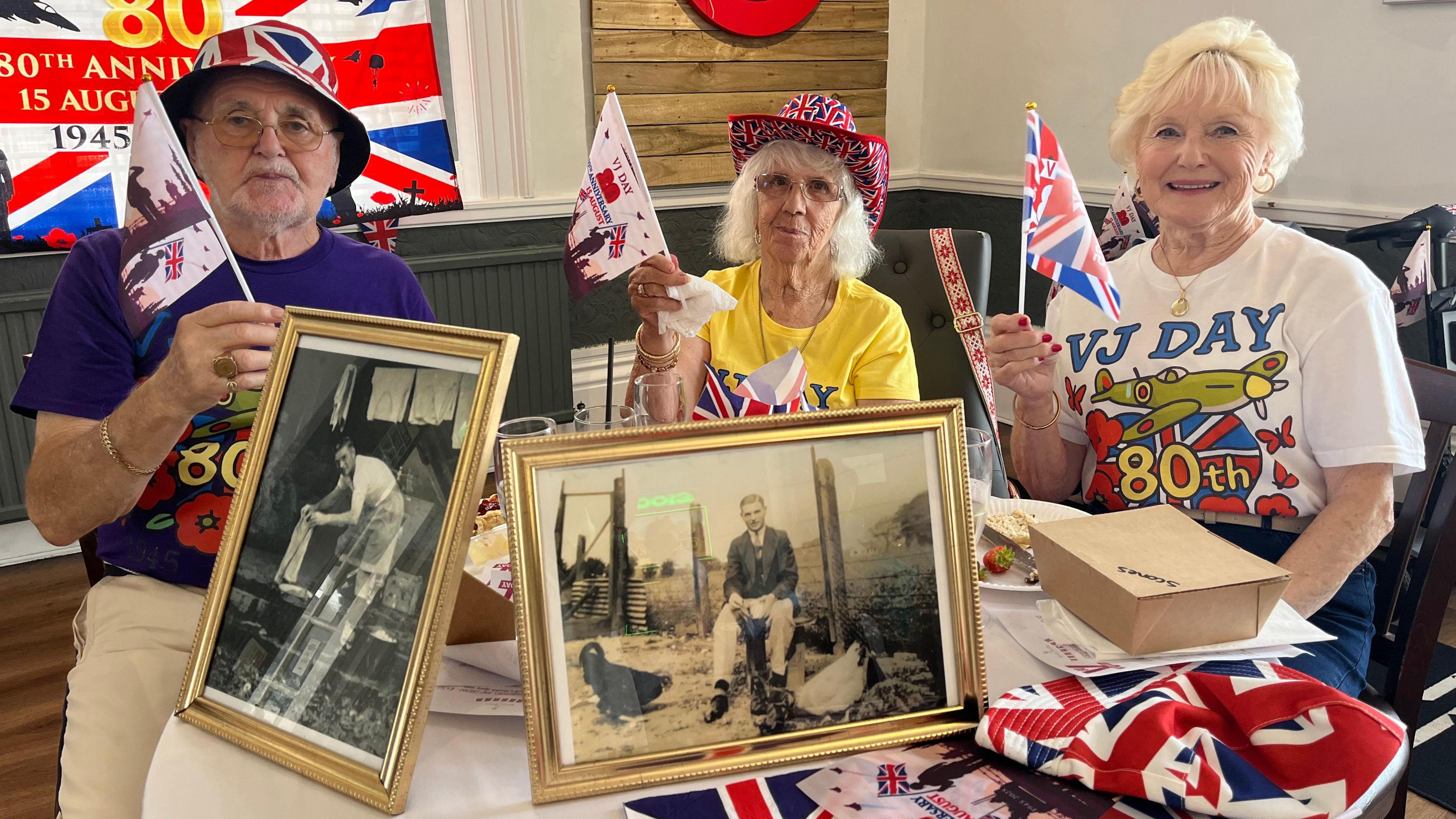 Three people sit at a table covered with old photos and British flags. A man sits to the left, holding a flag and wearing a union jack hat, and two women sit next to him wearing the same. 