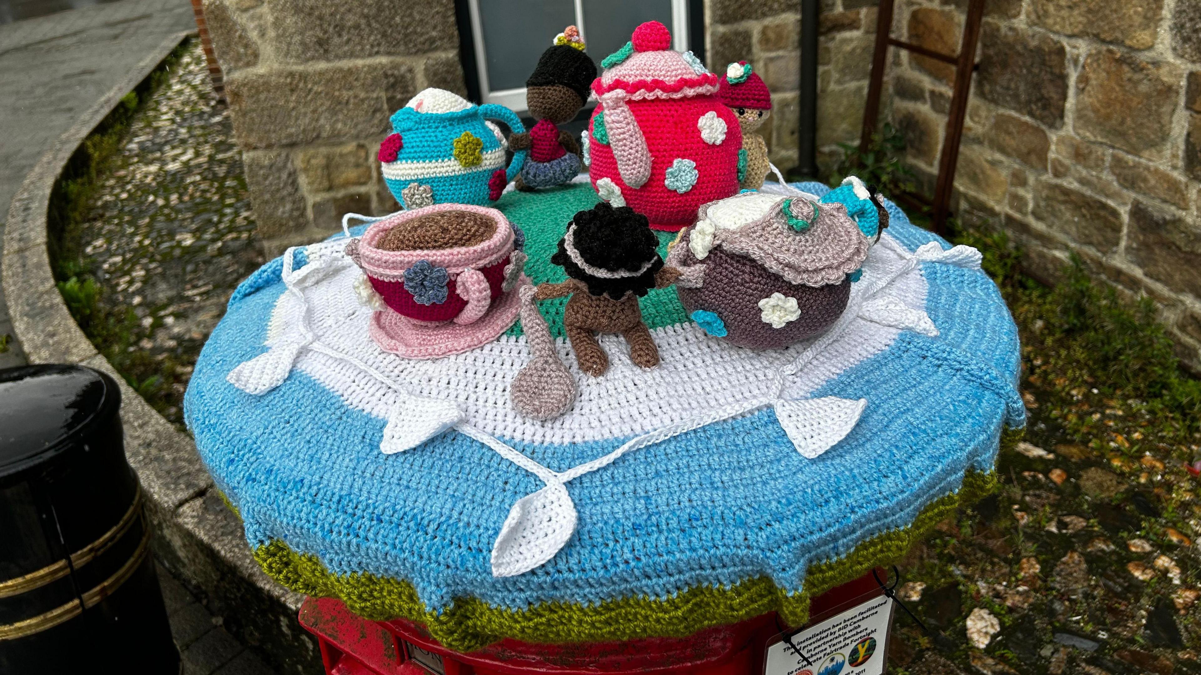 A blue and green postbox topper. There is a red and pink teapot with white, green and blue flowers on it, a brown sugar pot with flowers on it, a pink and red  mug and a blue and white milk jug, all knitted. There are knitter people also on the topper. 