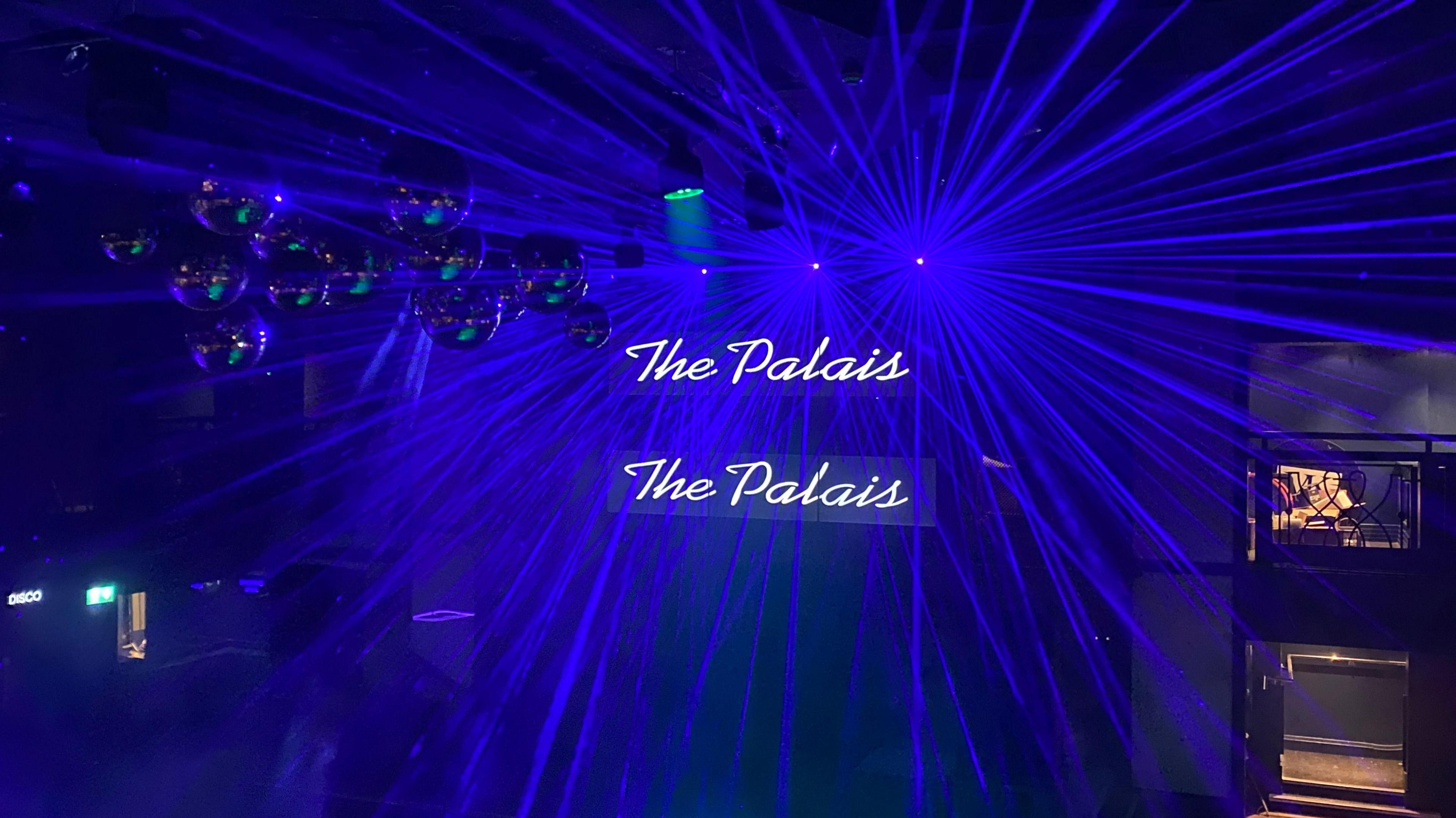 A dancefloor with two illuminated signs in the middle saying "The Palais", blue lasers of light surround the sign, and it is dark. 