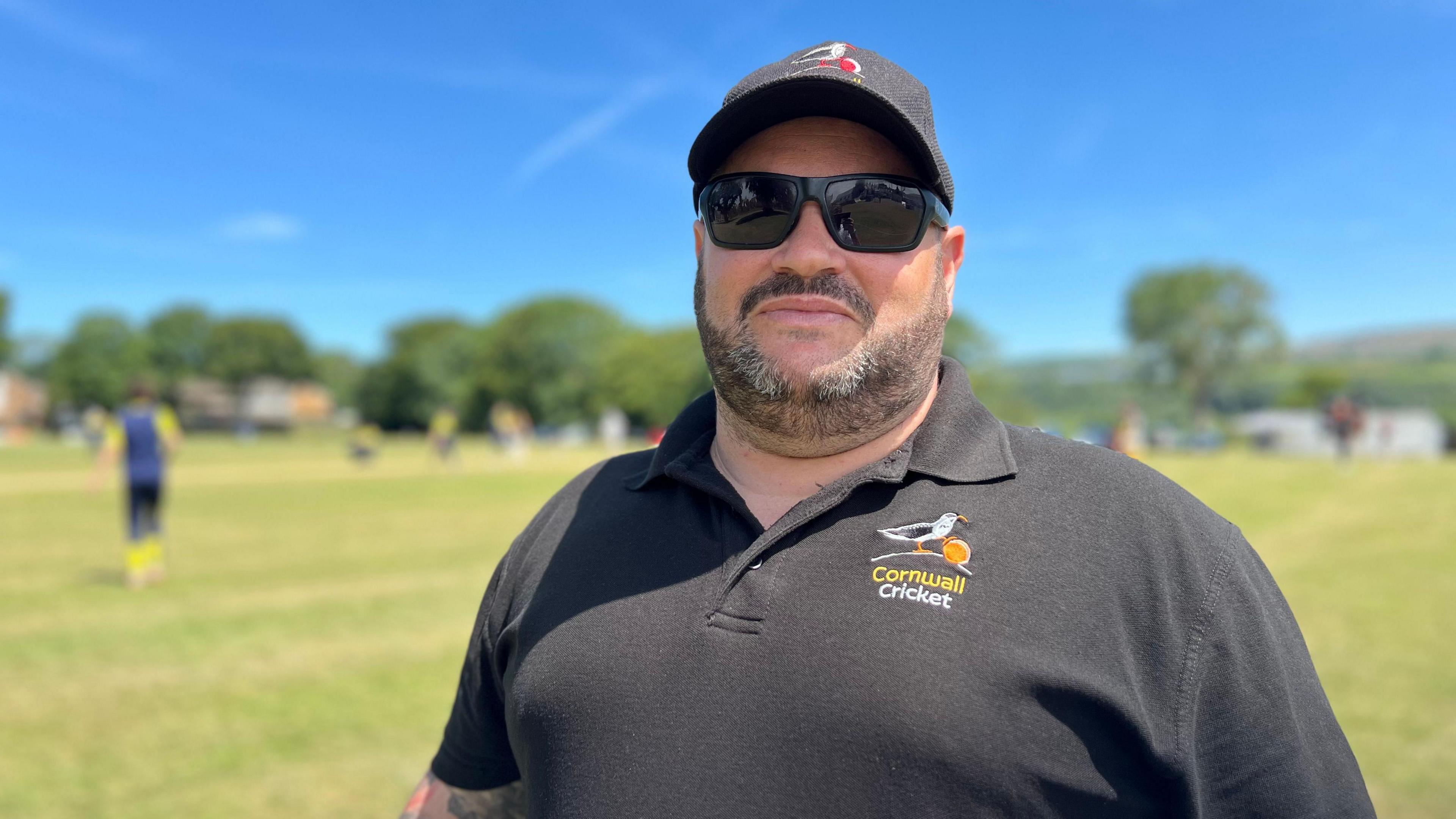 A large man wearing sunglasses and a polo shirt and a baseball cap looks at the camera 
