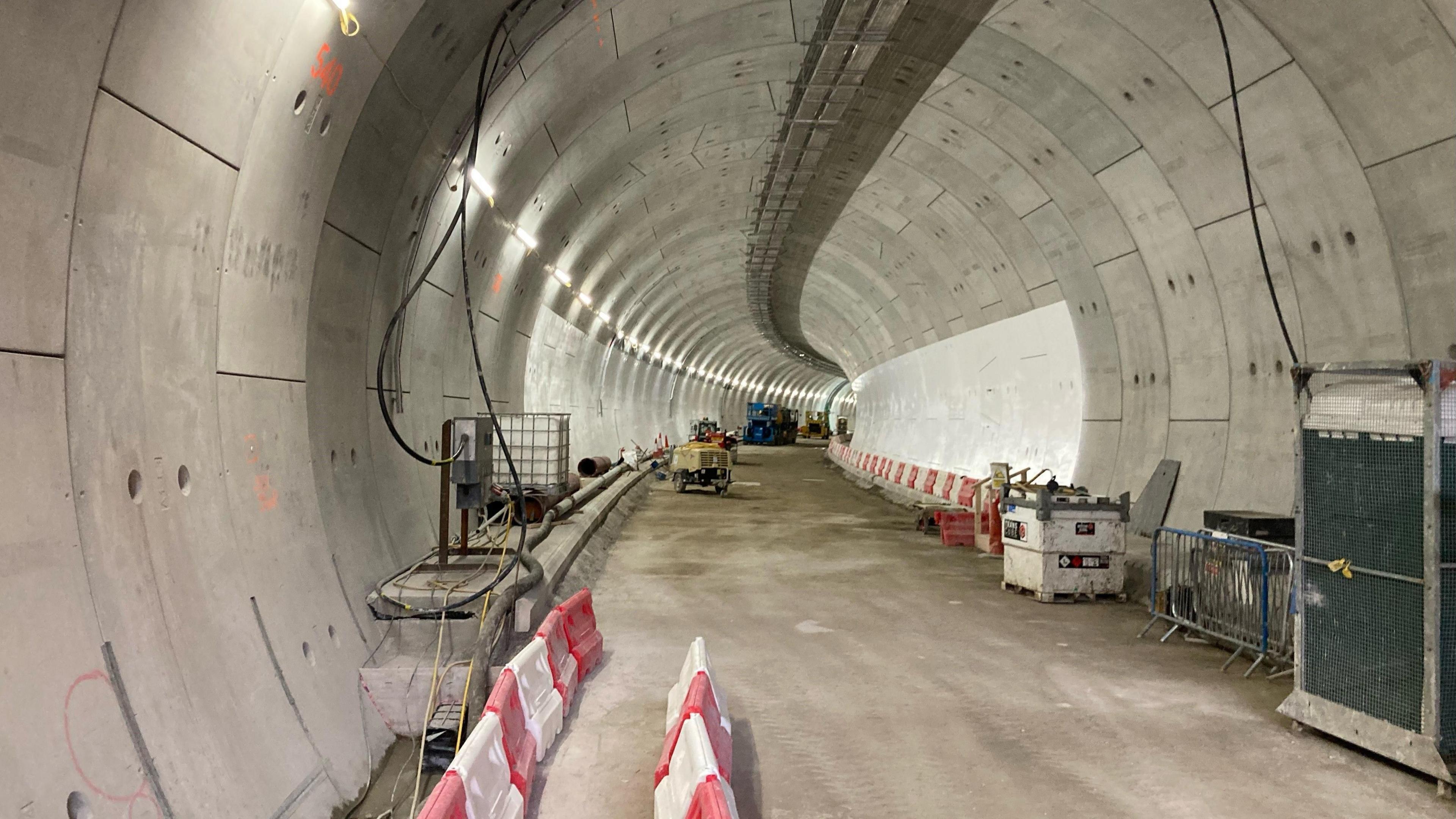 Inside construction of the Silvertown tunnel. The view shows the lit up inside concrete tunnel, with some red and white barriers on both sides of the tunnel and black wires hanging from the ceiling.