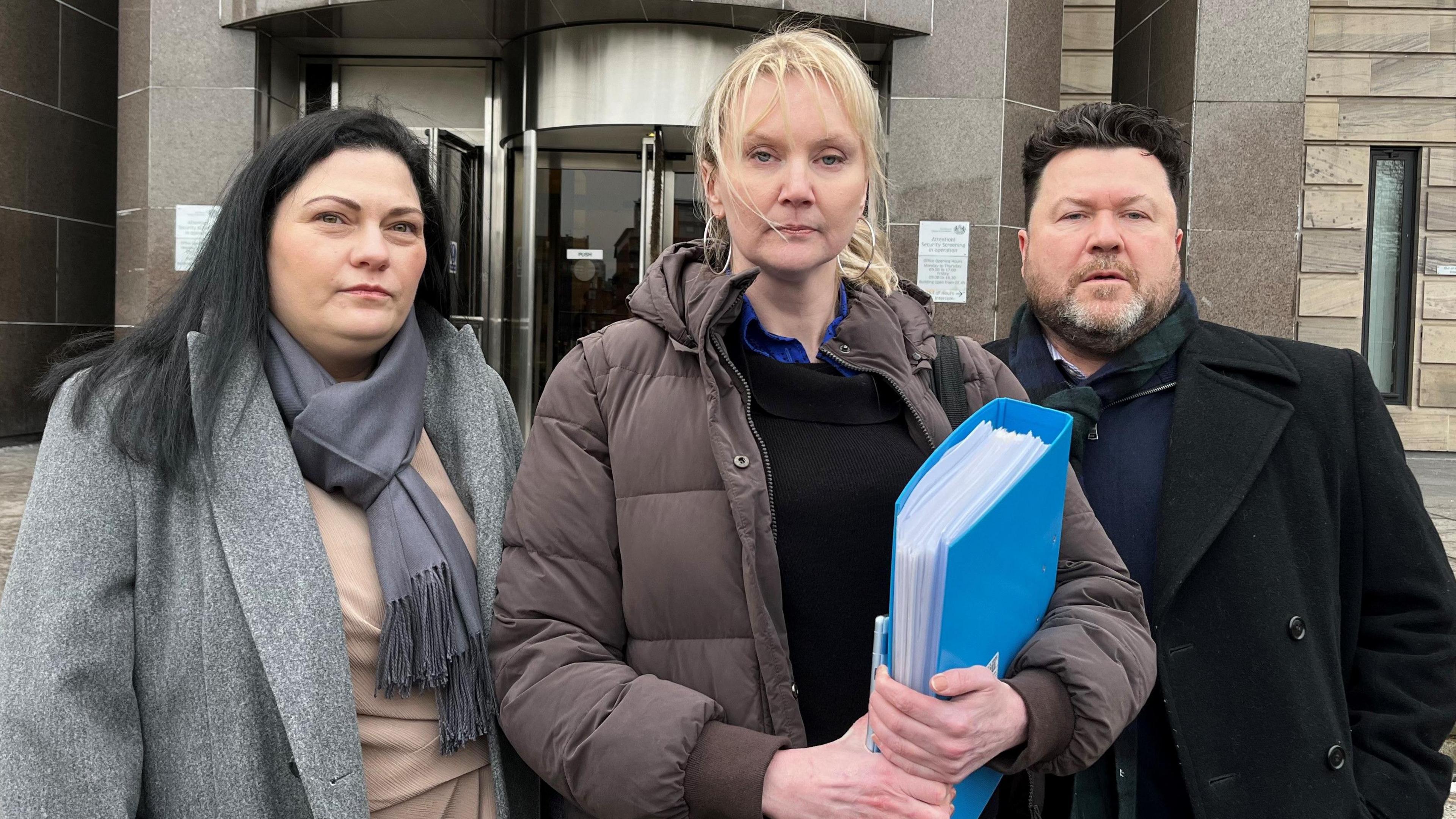 Theresa Smith is on the left. She has long dark hair and is wearing a grey scarf, grey woolen coat and a beige jumper. Their lawyer, a tall blonde woman, is wearing a brown puffer jacket, black jumper and blue shirt and is carrying a blue folder. She stands in the middle. Matthew Smith is wearing a black coat, dark blue cardigan and has a beard and greying black hair. All three are looking at the camera.