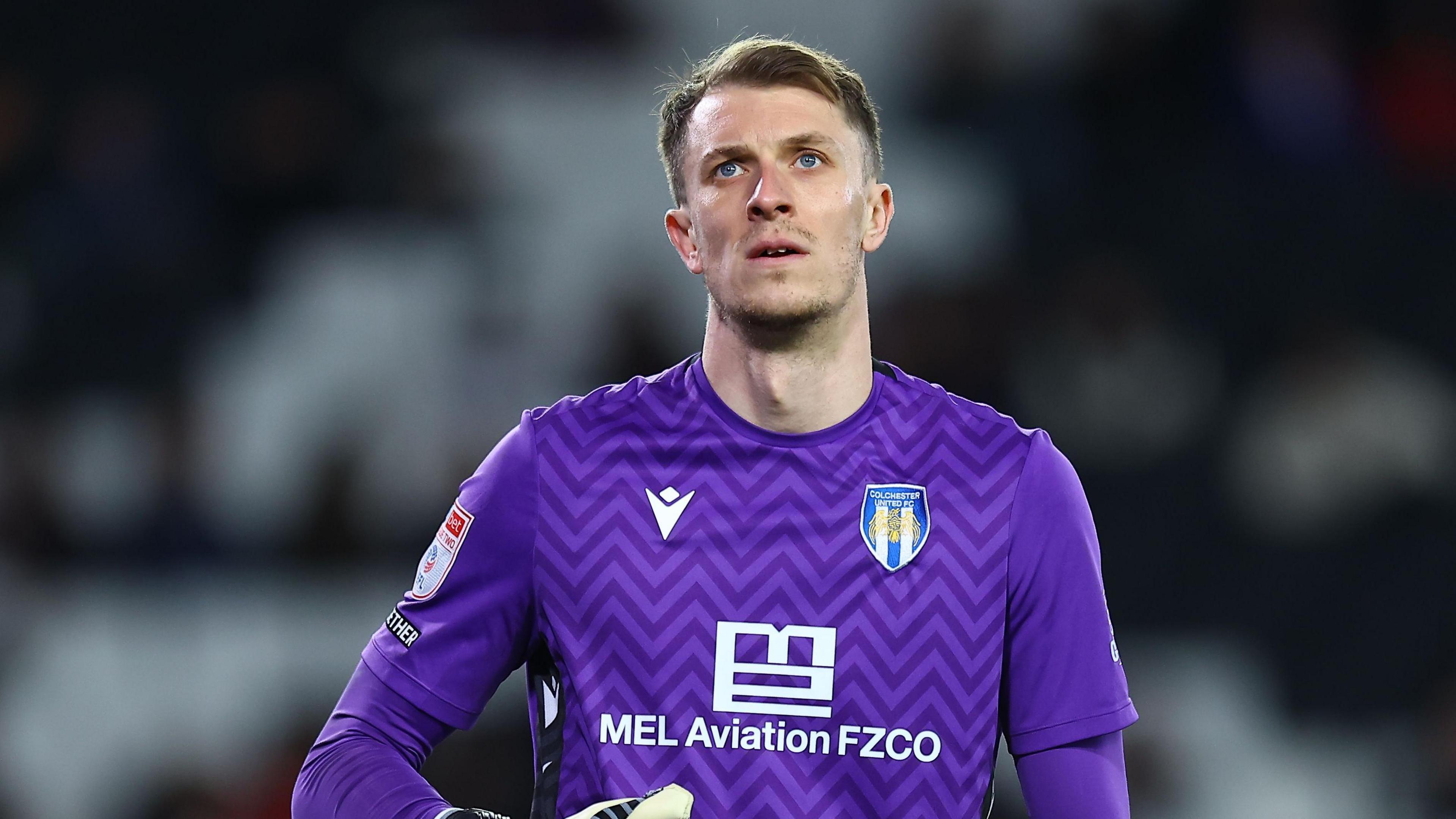 Matt Macey, wearing a purple Colchester United goalkeeper kit, looks upwards during a game