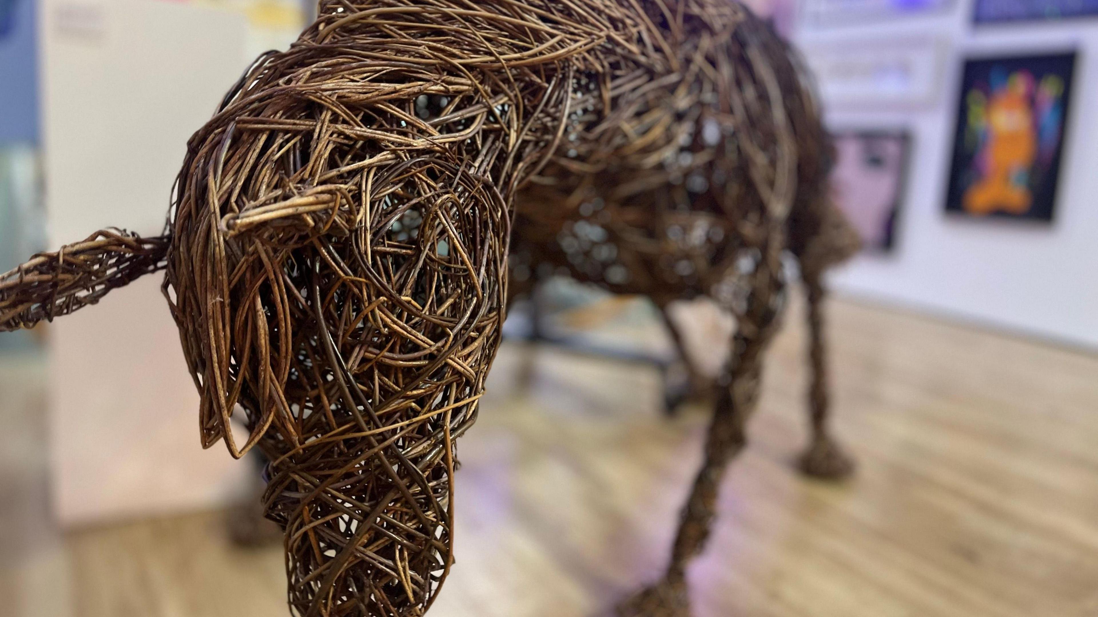 A near-life-size wicker sculpture of a horse in a gallery with paintings behing.