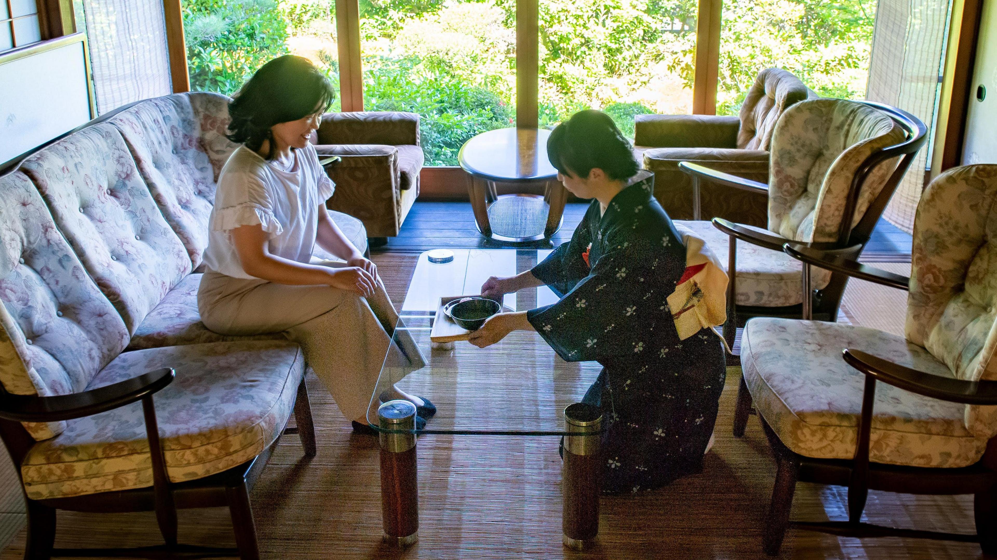 A photo of a traditional Japanese tea ceremony. A woman on the left sits on a couch as a host, dressed in a kimono serves her a cup of matcha. 