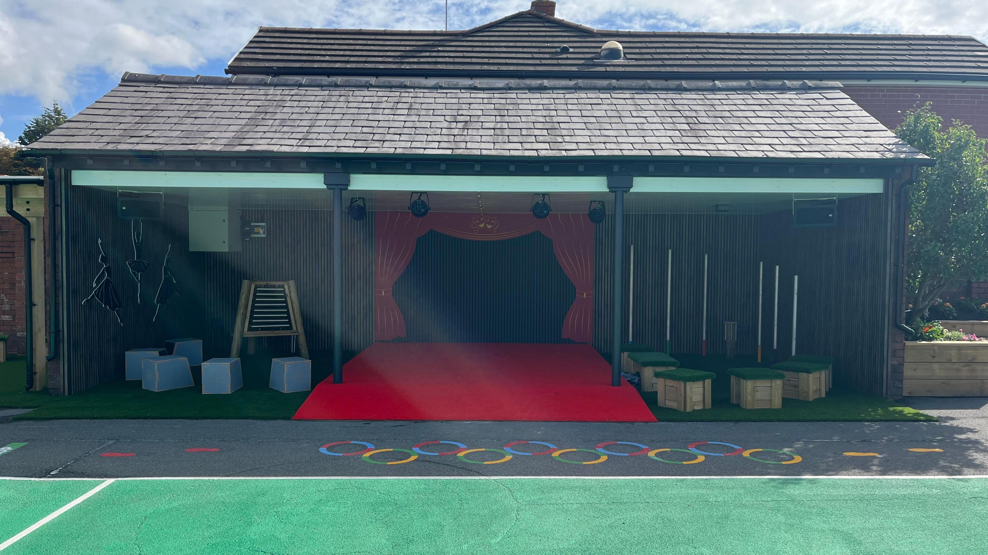 A red stage with red curtains situated under the roof of an open-side building.