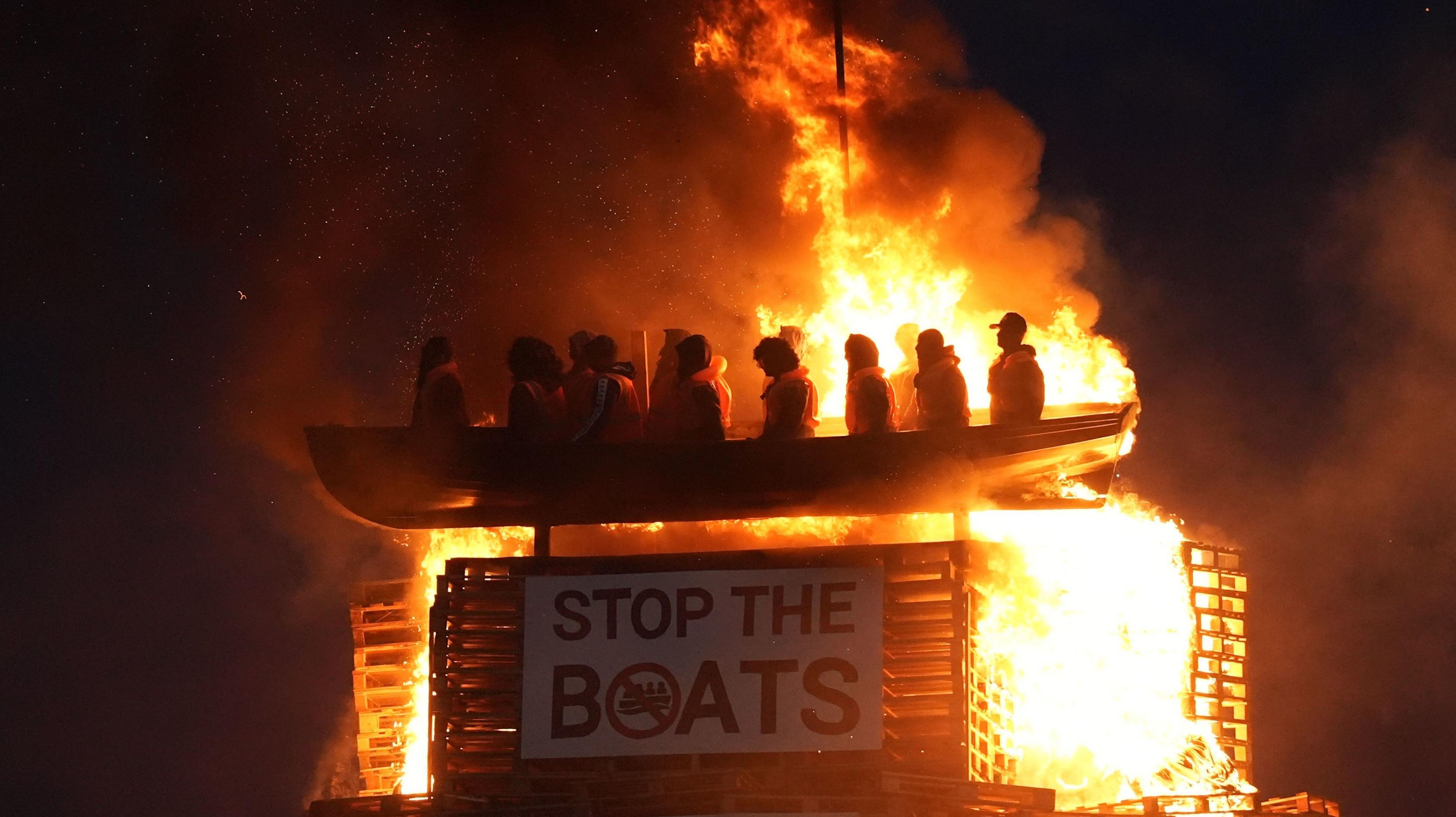 The picture shows a model of a small boat with figures inside in orange fluorescent vests. Below is a sign which says 'STOP THE BOATS'. It is on fire.