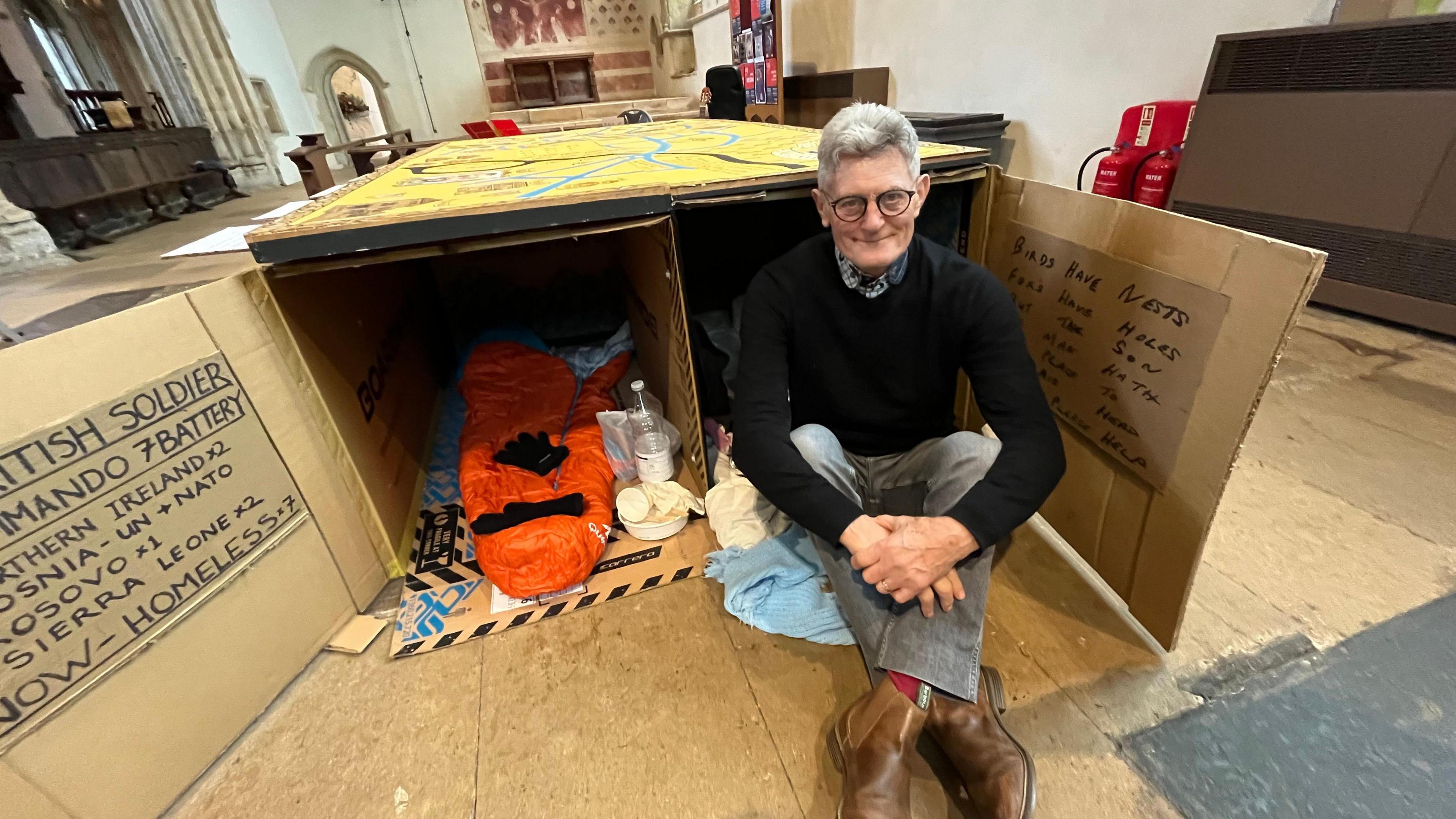 Adrian Brooks, who is wearing grey jeans, a black jumper and glasses sat down cross legged in front of the artwork - which is made of cardboard boxes and contains discarded food wrappers.