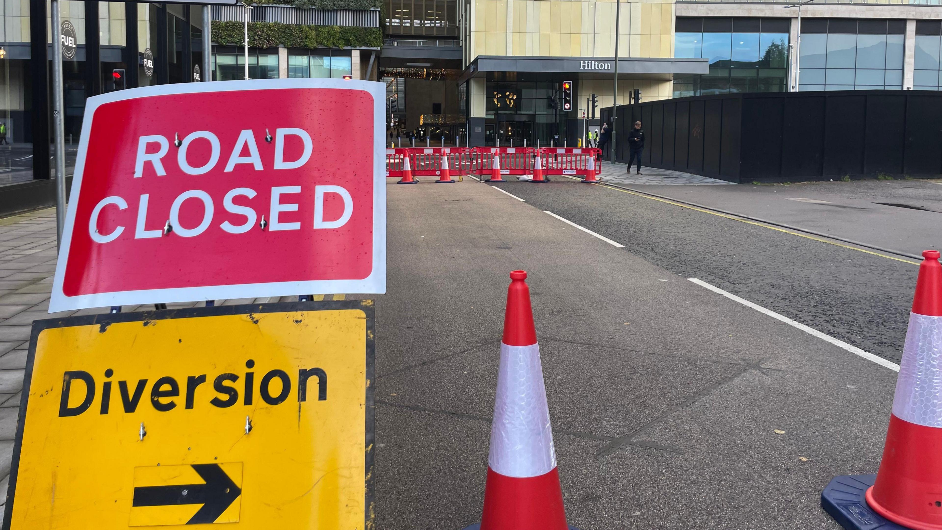 A road closed sign with a diversion arrow underneath it. There are cones also in shot and barriers seen behind. Further in the distance is the front entrance to the Hilton Hotel 