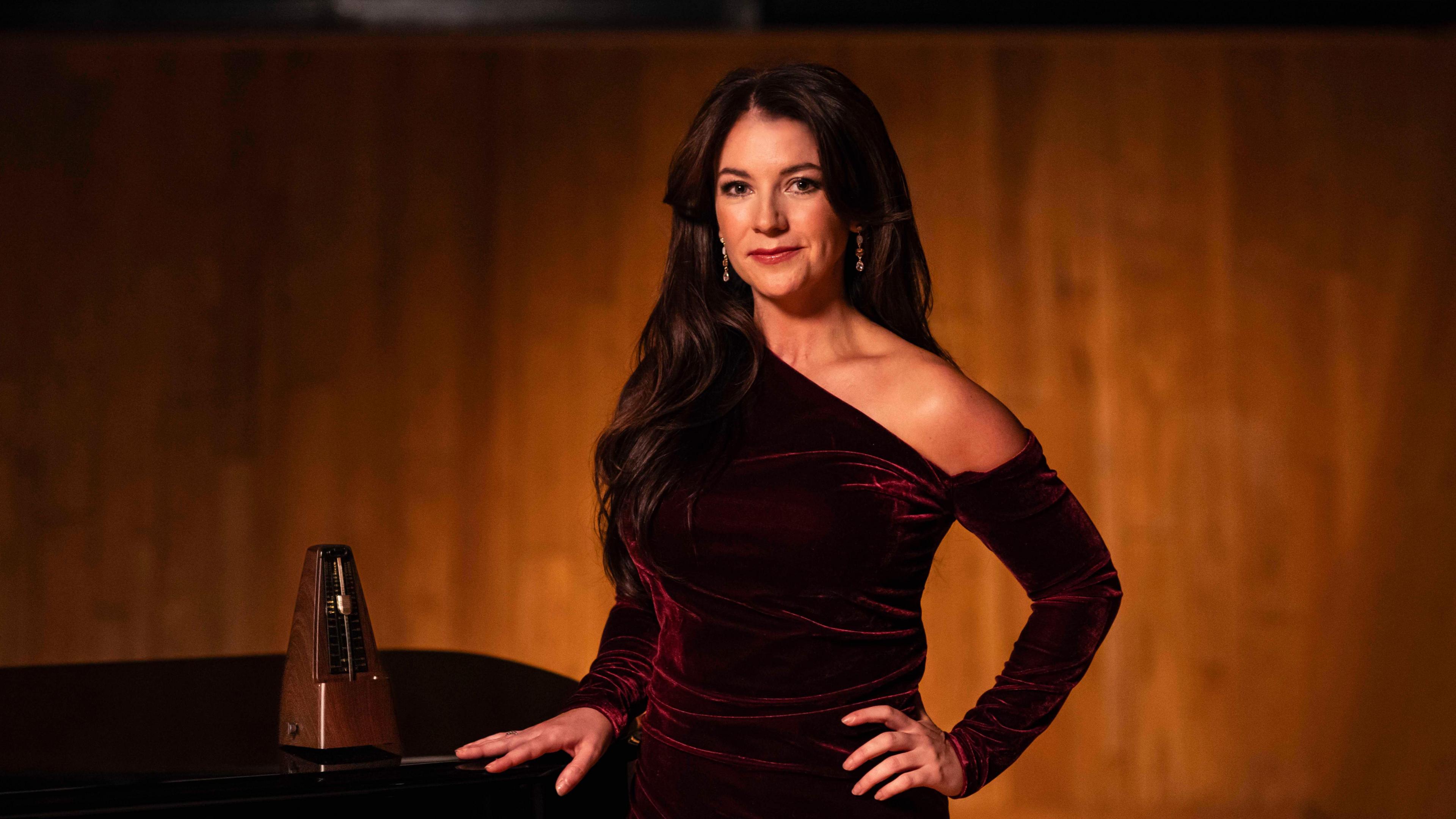 Monica McGhee, a woman with long dark hair wearing a dark red velvet dress, stands to the right of the piano with her hand on it. A wooden metronome can be seen beside her