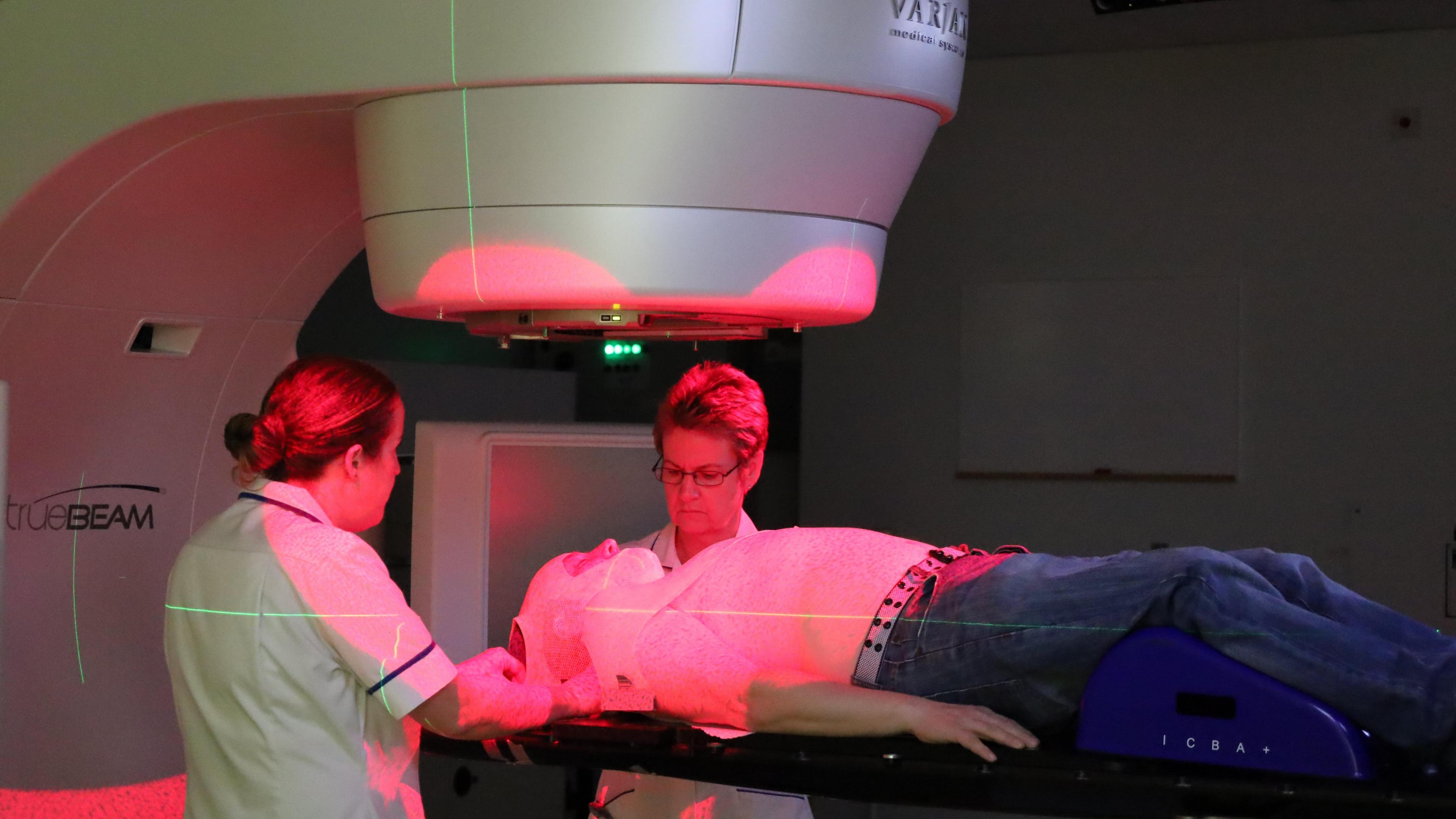 A cancer patient undergoes radiotherapy treatment. They are surrounded by two nurses and are bathed in red and green lights. The patient is wearing a face mask.