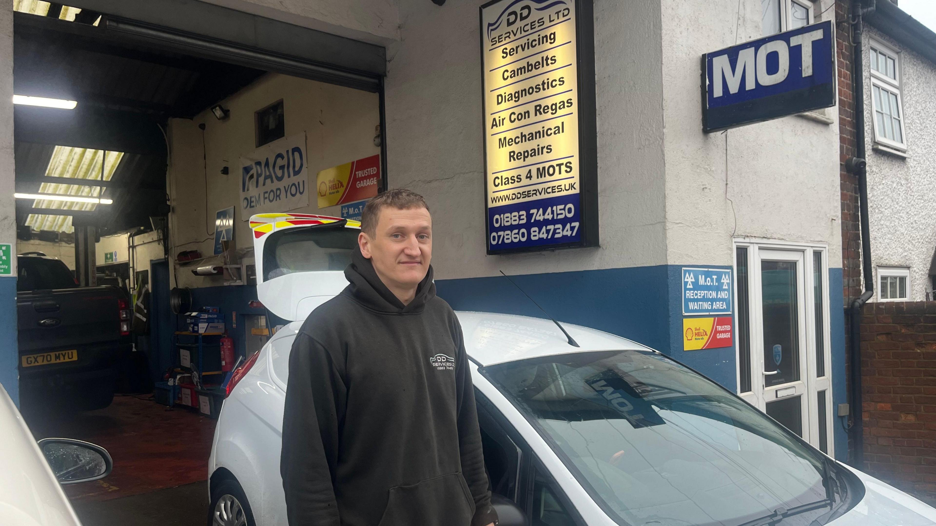 Shane Fry stands in front of a white hatchback car in the entrance to his garage in Godstone. He wears a dark branded fleece with DD Motors embossed on it.