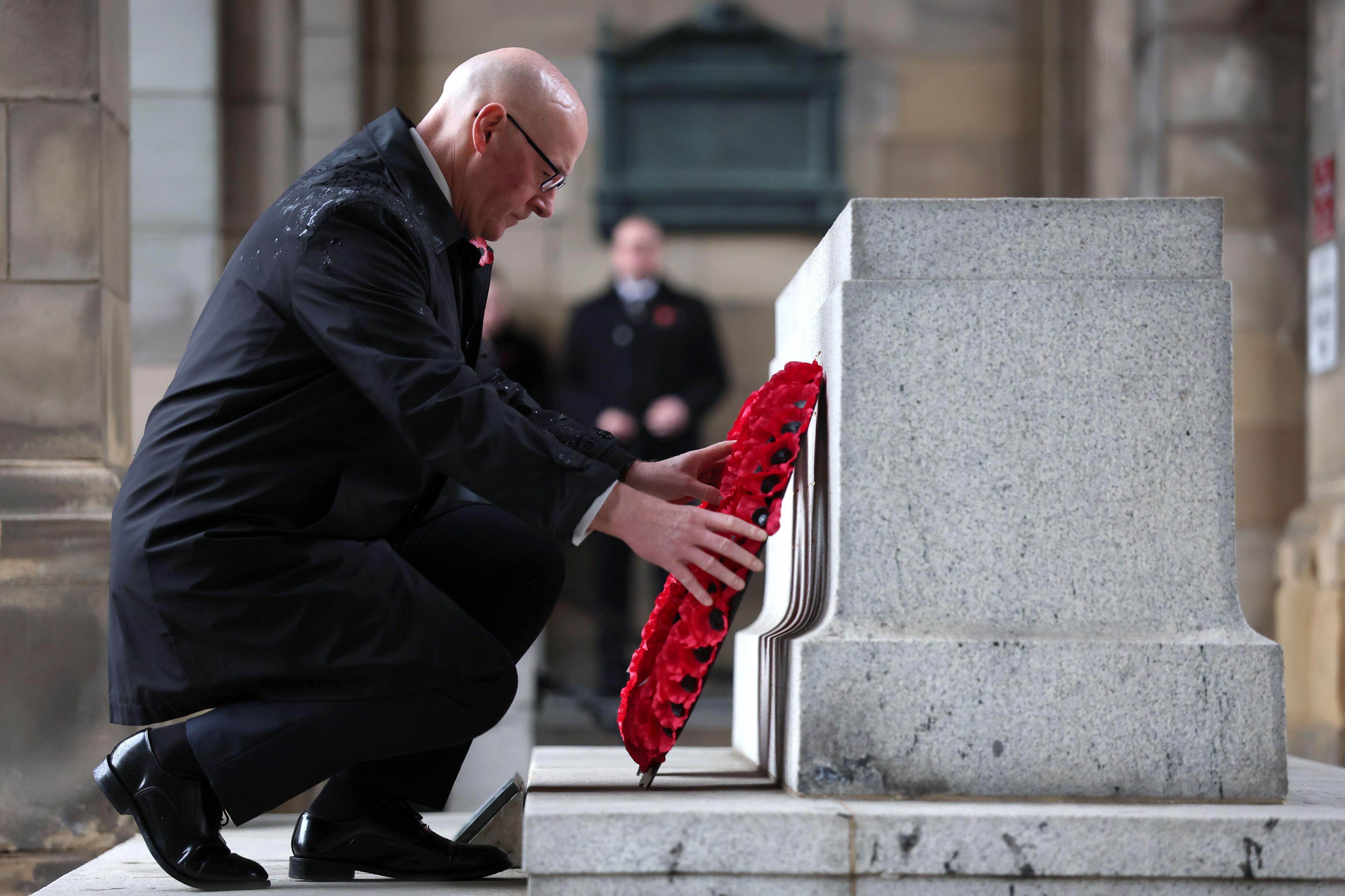 John Swinney crouches and lays a red poppy wreath at the Stone of Remembrance at the entrance to Edinburgh City Chambers on the Royal Mile. He has a black raincoat and highly polished black shoes.