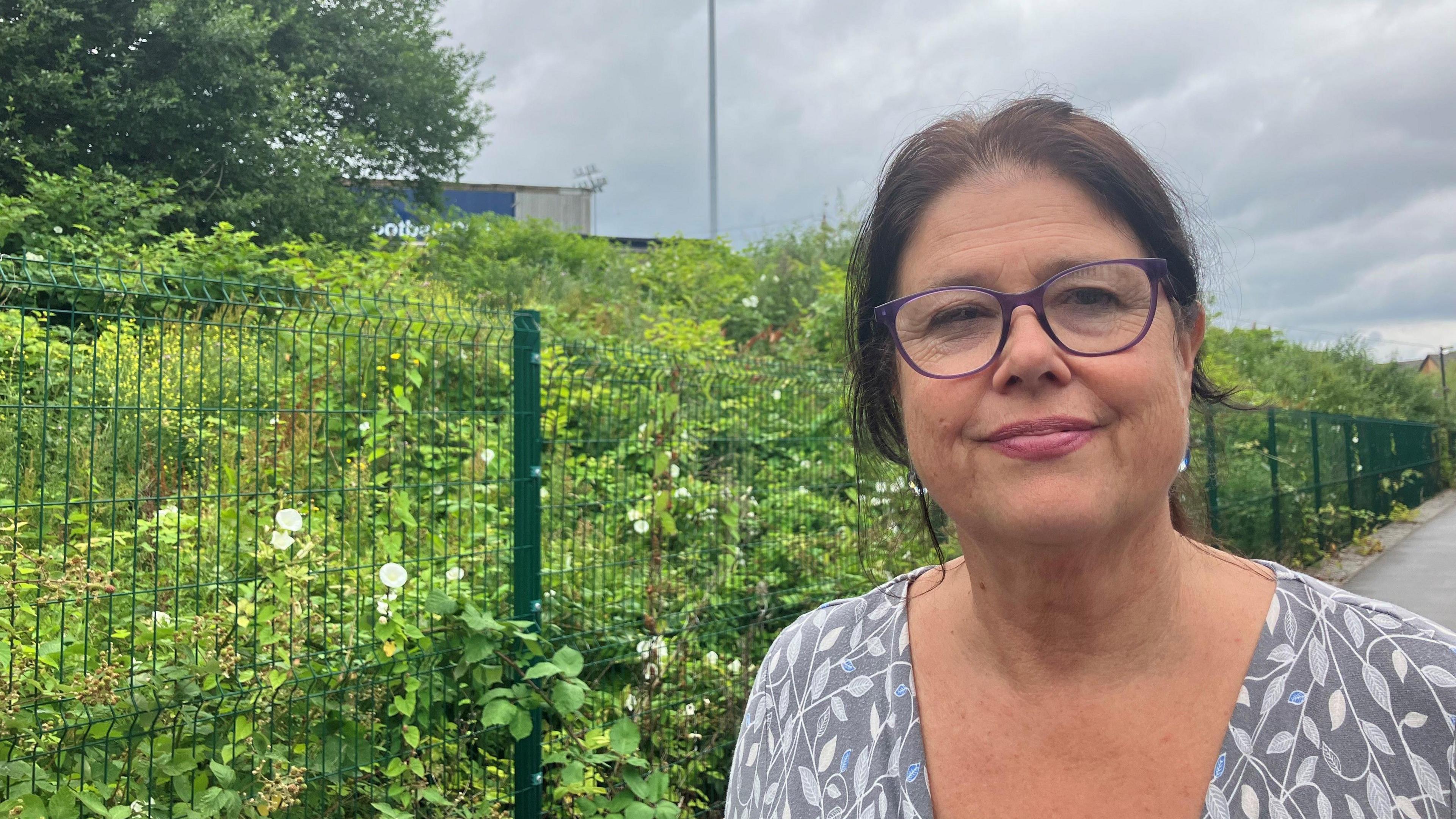 Photograph of Julia Blundell who lives near Edgeley Park. She is standing in front of a fence and the Edgeley Park football stadium is in the background.