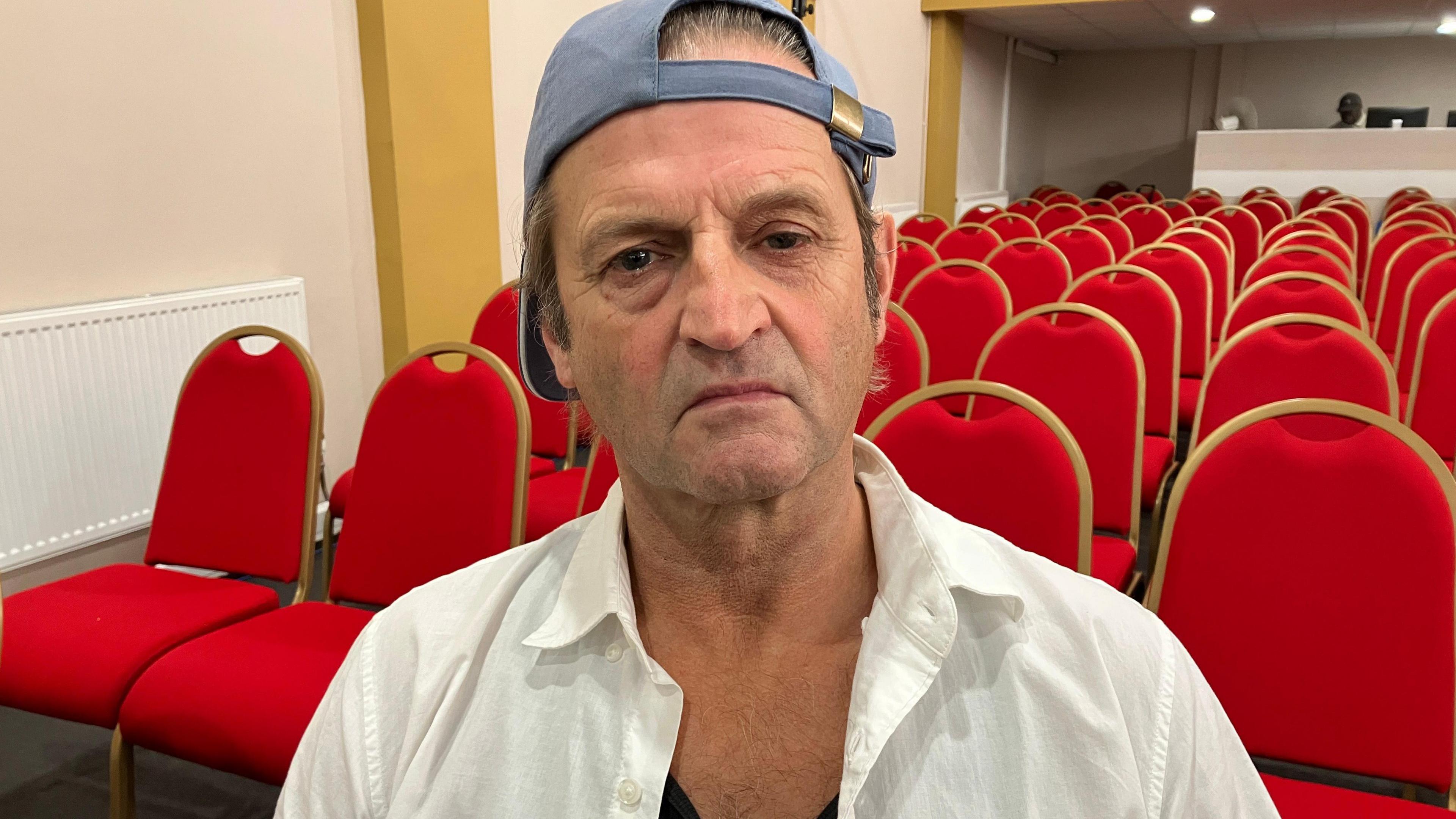 A man wearing a white shirt and a light blue cap looks into the camera. Rows of red chairs are in the background.