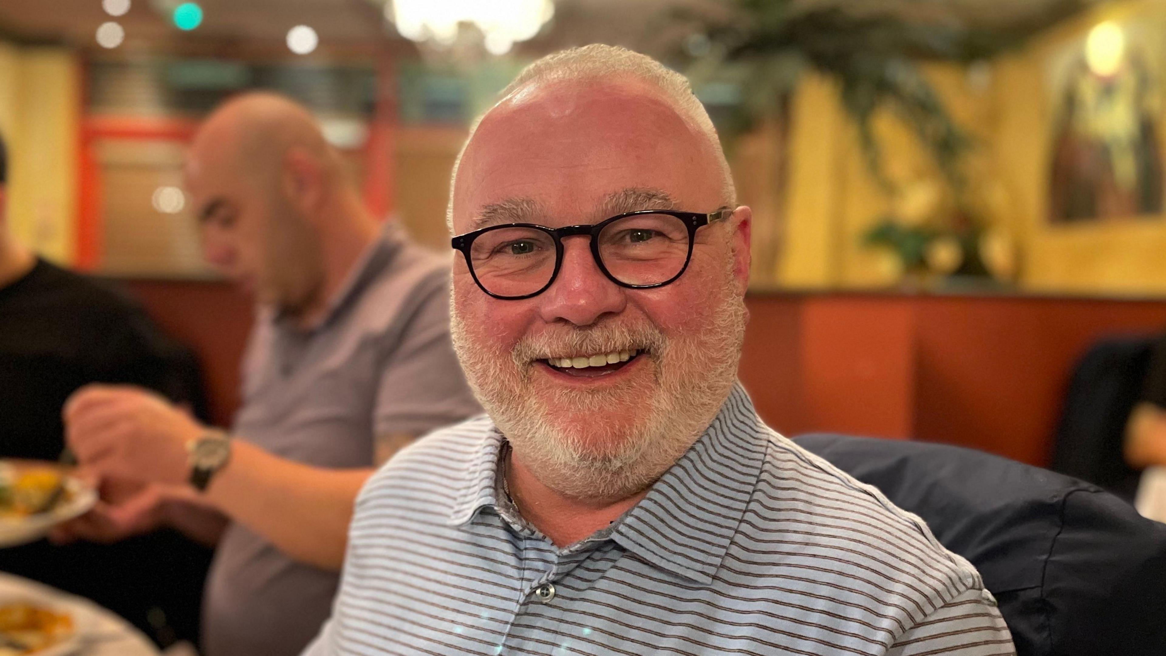 Wayne Fitzgerald smiling and looking direct to camera. He is wearing black-rimmed glasses, a horizontally striped t-shirt, and has a short, grey beard. Other diners can be seen out of focus in the background.