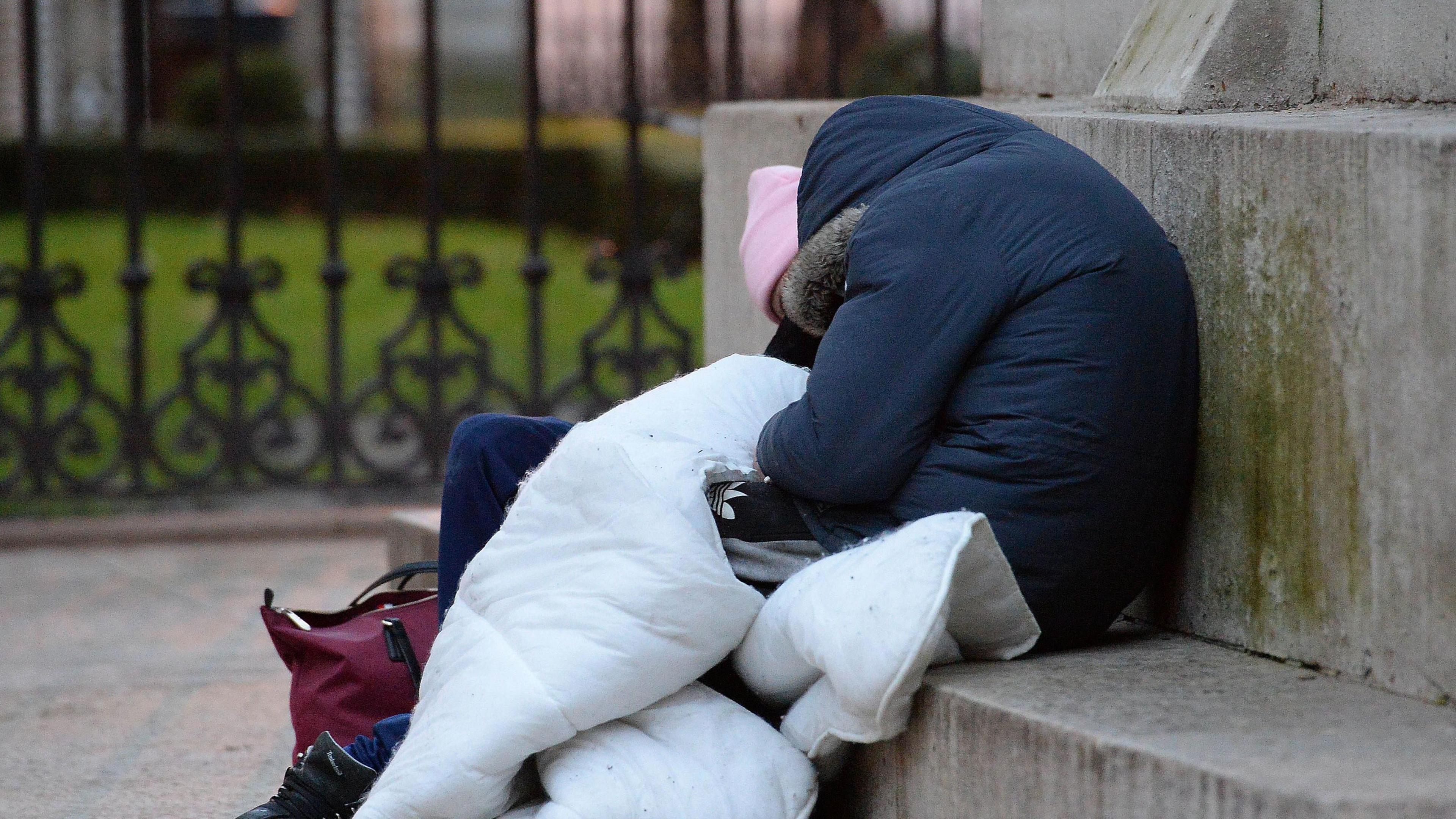 A rough sleeper wrapped in a sleeping bag is sitting on a concrete step and is slumped over