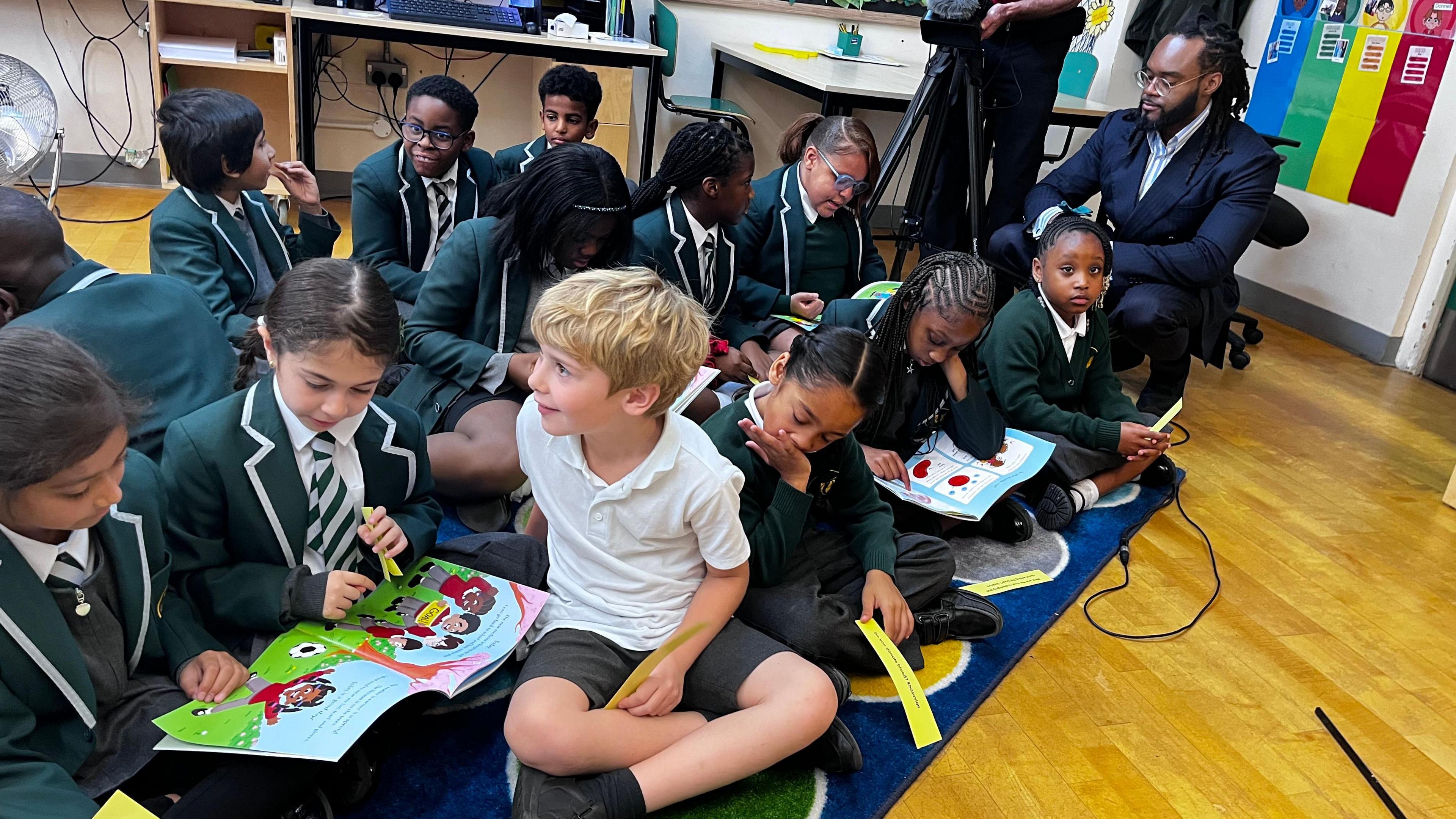 A group of primary schoolchildren sat on the wooden floor of a classroom. They are all dressed in school uniform, wearing white shirts and green jumpers or blazers. They are reading copies of the 'My Blood, Your Blood' book, with colourful illustrations visible.