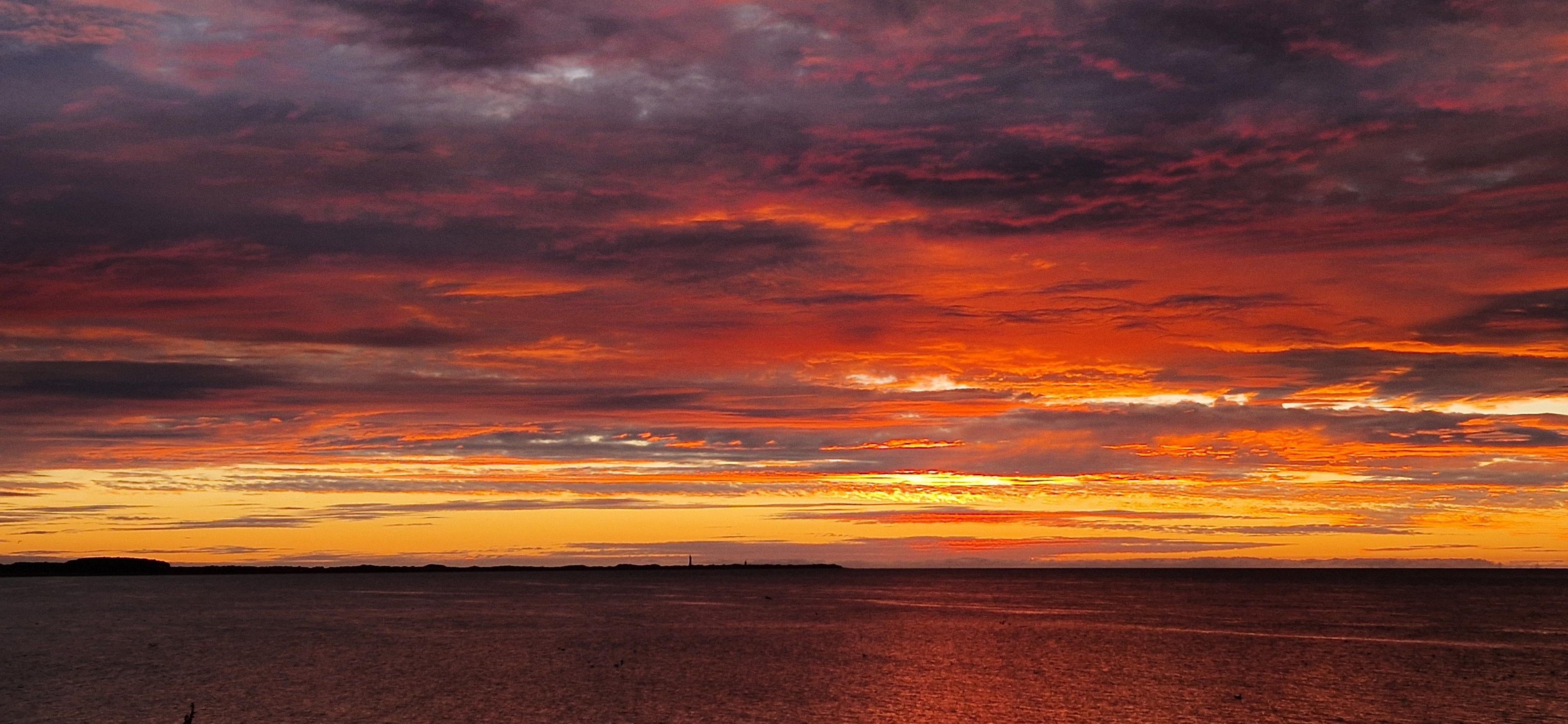 A vivid sunrise paints the sky in fiery shades of orange, red, and purple, blending into darker clouds above. There is a calm sea below reflecting the sunset.