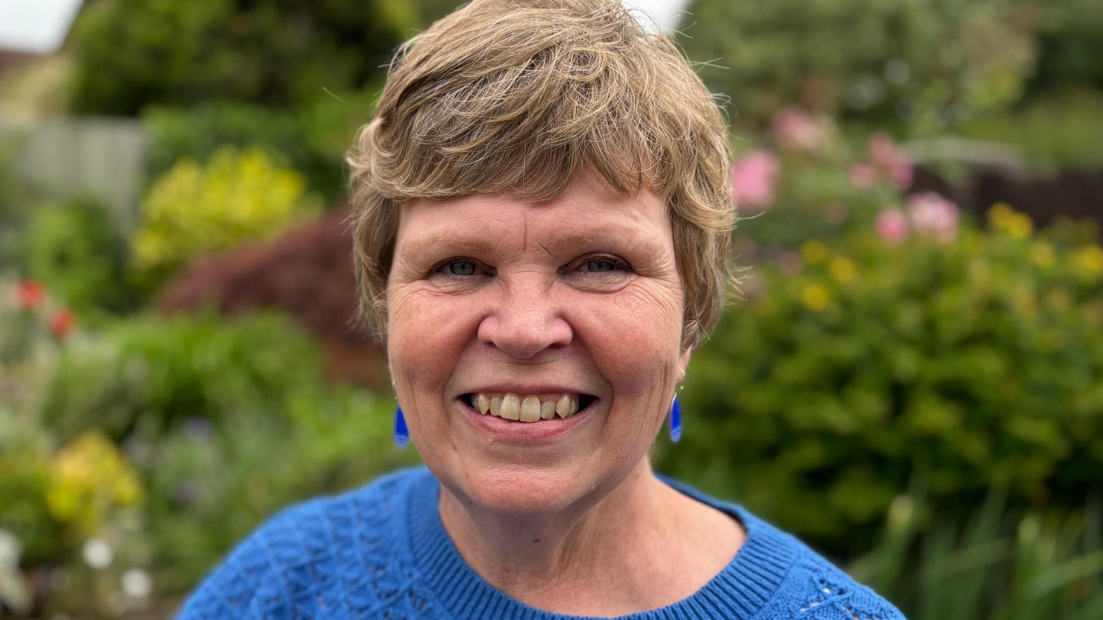 Gwen, who has short blonde hair, is looking at the camera and smiling. She's wearing a blue crew-neck jumper and she has blue earrings. She's standing in a garden which is out of focus behind her. There are many green plants and bushes.