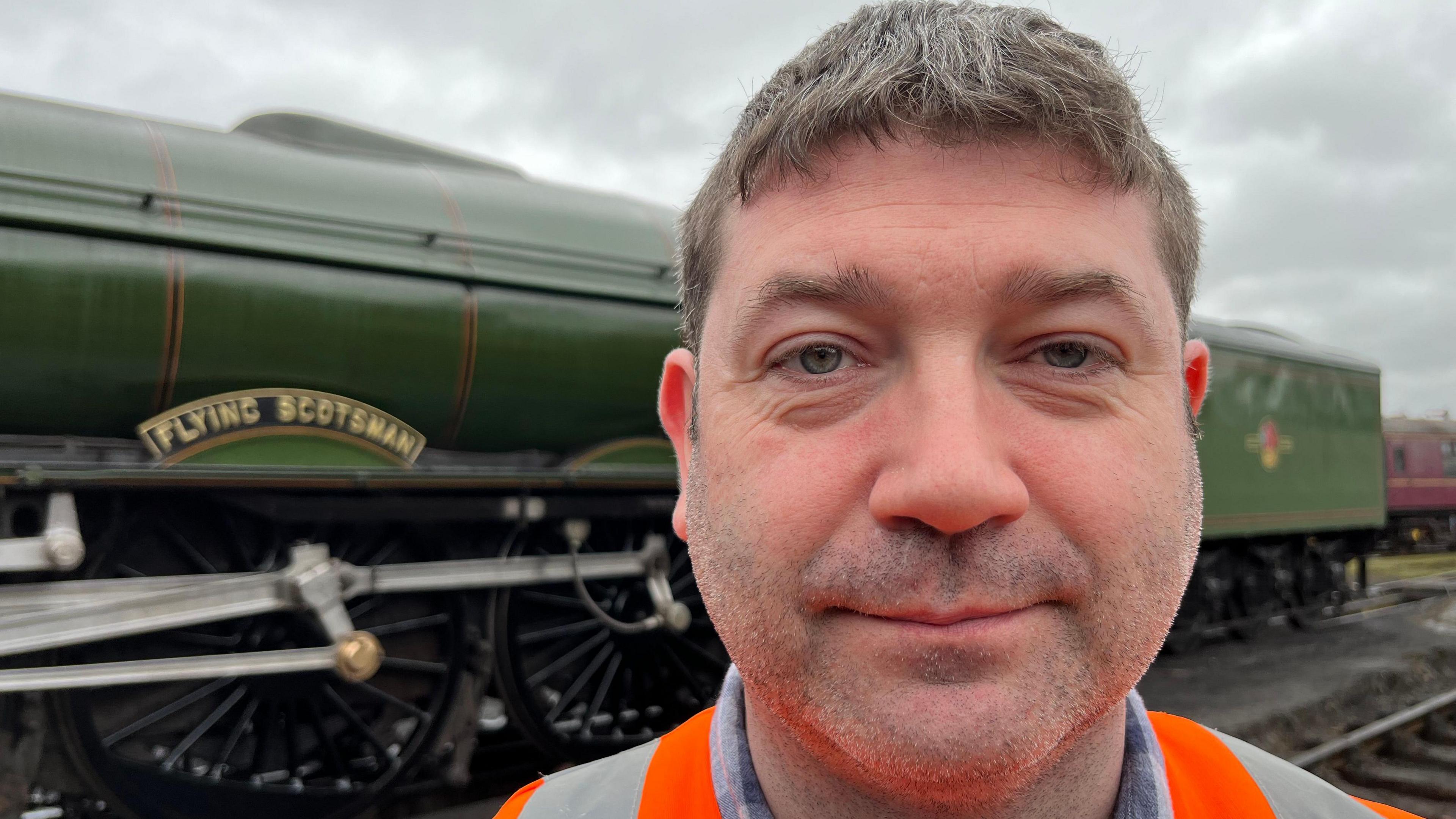 A smiling man with greying hair. Wearing an orange and grey high-visibility jacket - standing in front of the Flying Scotsman. The famous steam locomotive is green with glossy black wheels. 