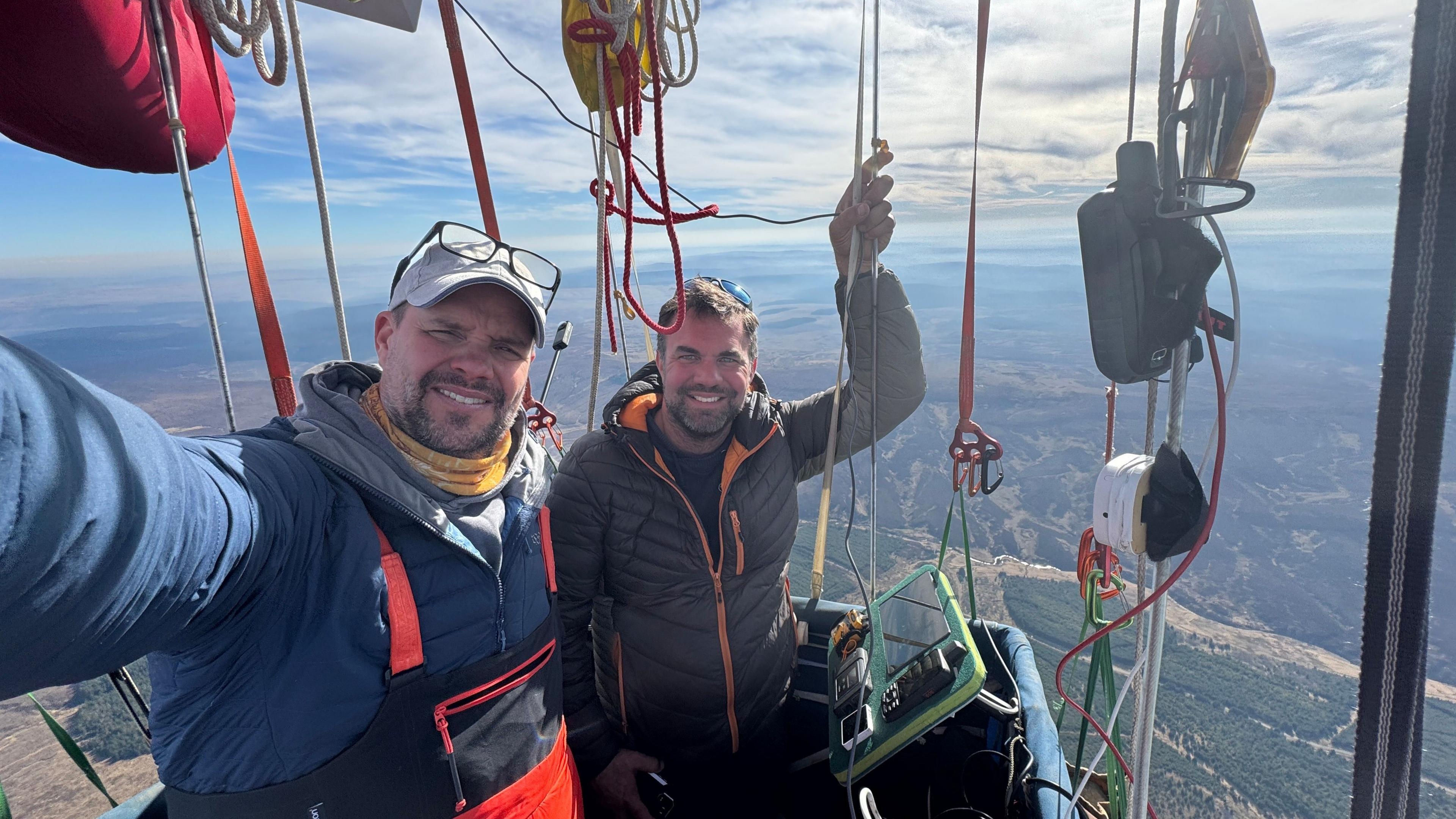 Two men standing in the basket of a hydrogen balloon, smiling straight at the camera. The balloon is in flight and the British countryside can be seen in the background.