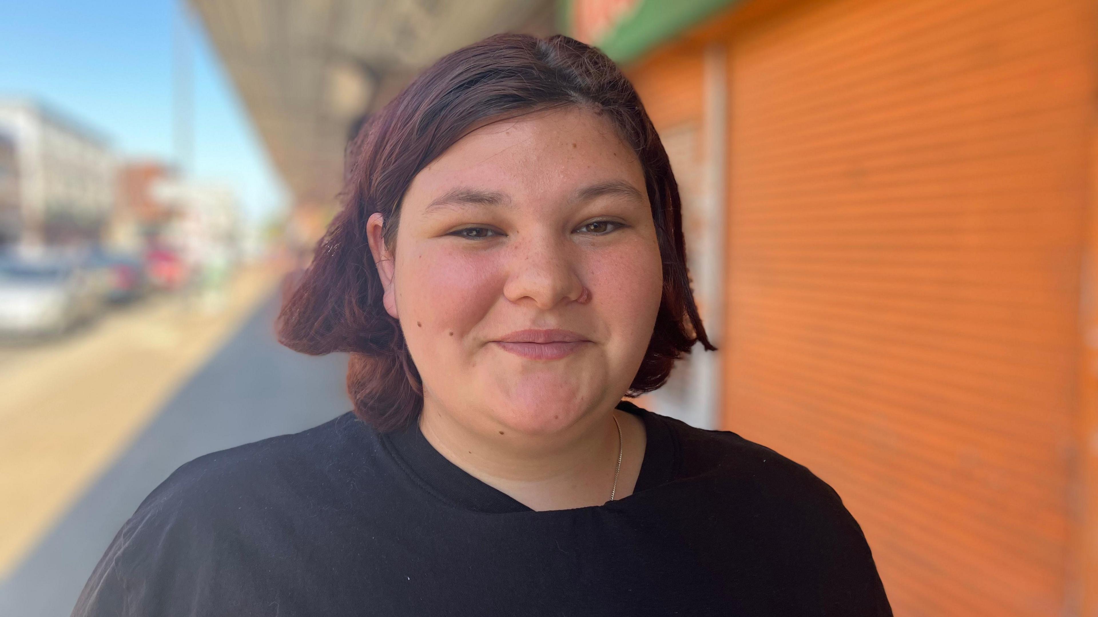 Libby smiles into the camera in front of a row of shuttered shops on a sunny day in Grimsby. She has cropped red hair and is wearing a black top.