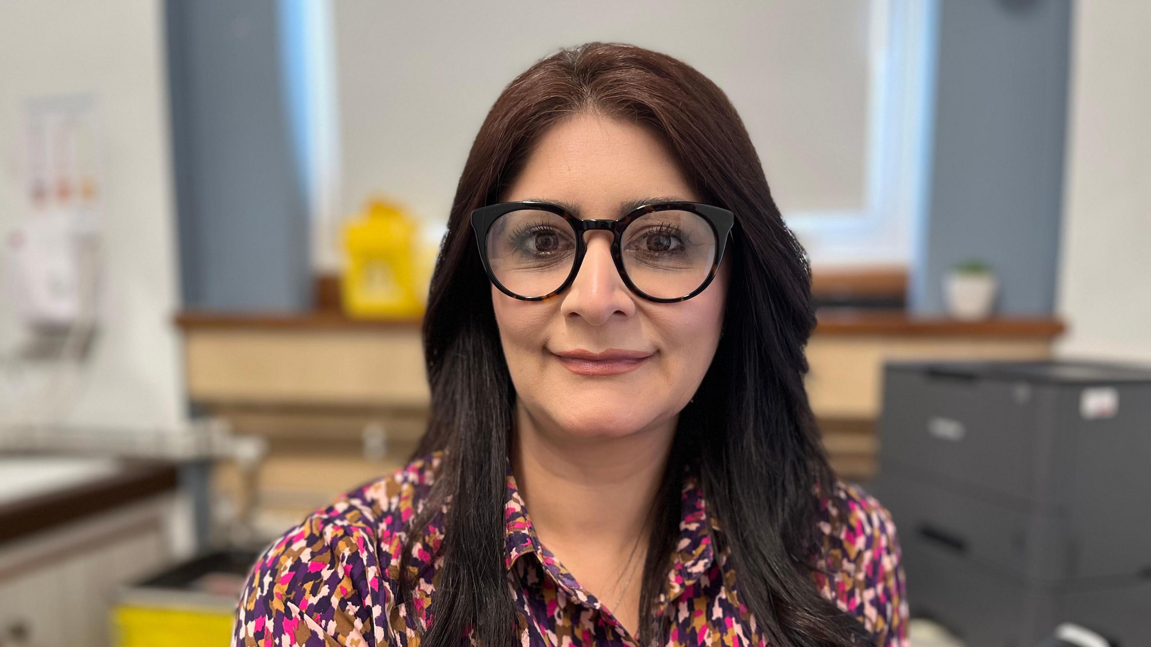 Shabina Qayyum stares at the camera. She wears black rimmed glasses and wears a colourful patterned top. In the background is a clinical office.