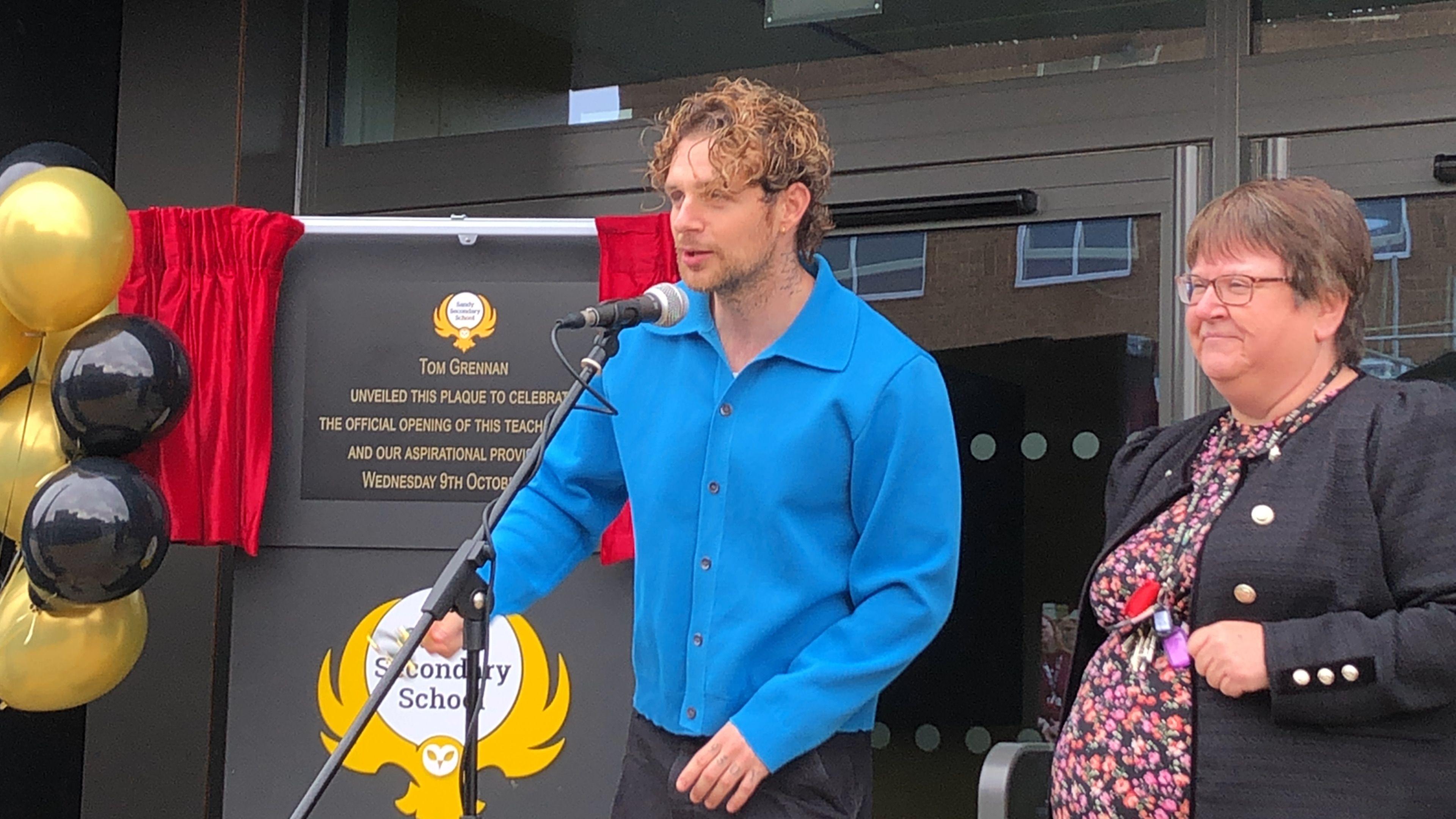 Tom Grennan pictured behind a microphone at Sandy Secondary School. He wears a bright blue long sleeve shirt with black buttons. He has curly hair, a gold ear piercing and a tattoo on his neck. On his right is Karen Hayward who has short brown hair, a black blazer with gold buttons and is wearing a floral dress. 