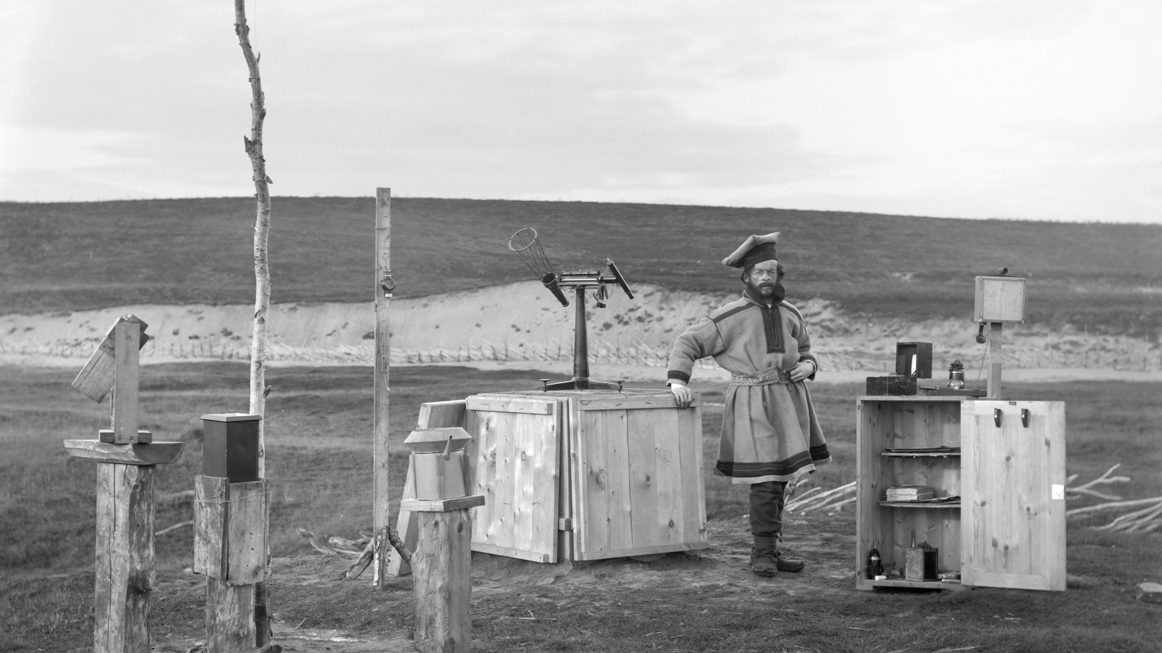 A self-portrait taken by Sophus Tromholt. He is posed amongst scientific equipment, dressed in Sami clothing - a hat, belted coat and knee-length boots.