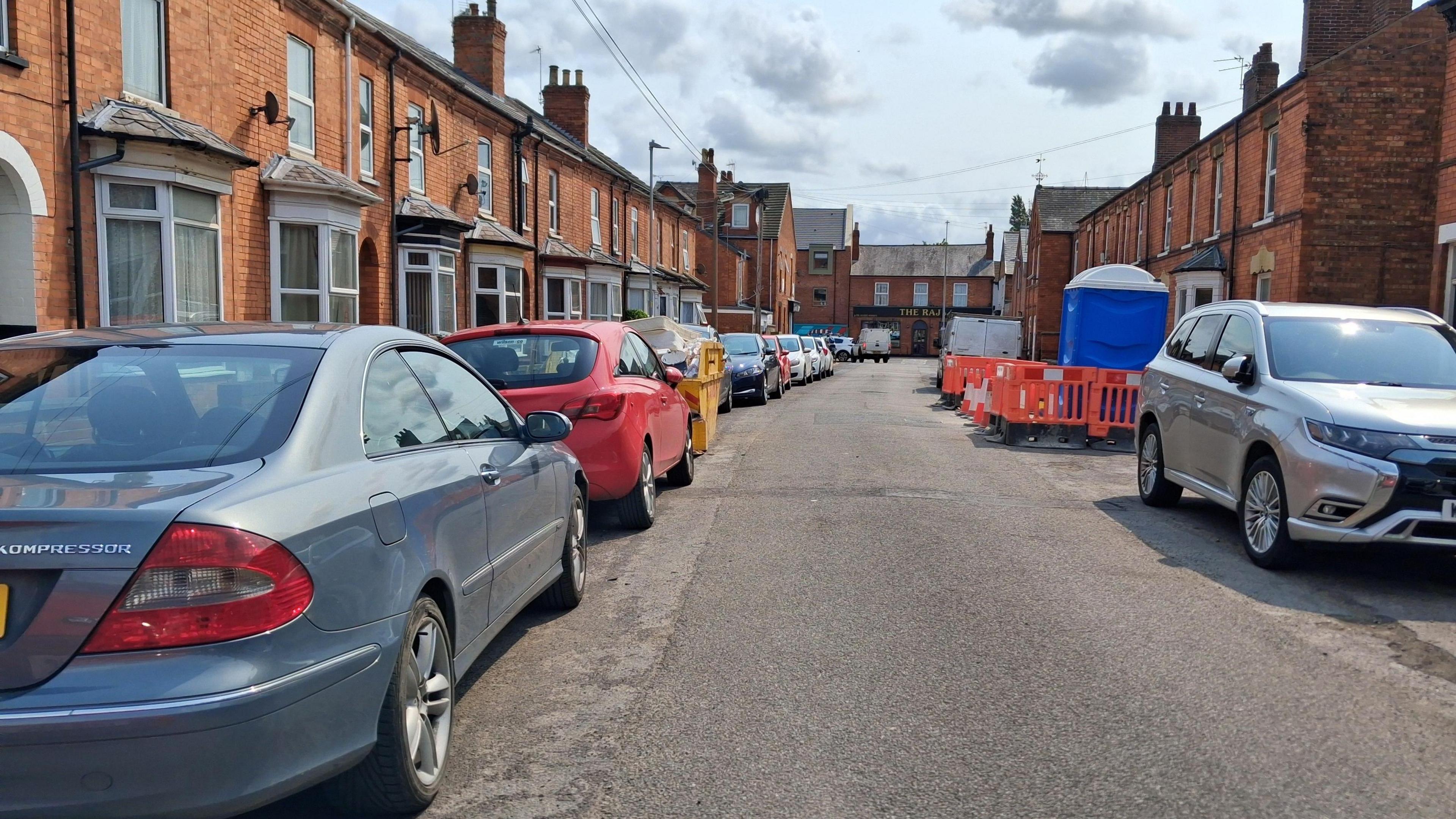 Cars parked on both sides of a residential street. There is a skip and and portaloo with a barrier around it in between some of the cars. It is a bright day with some clouds.
