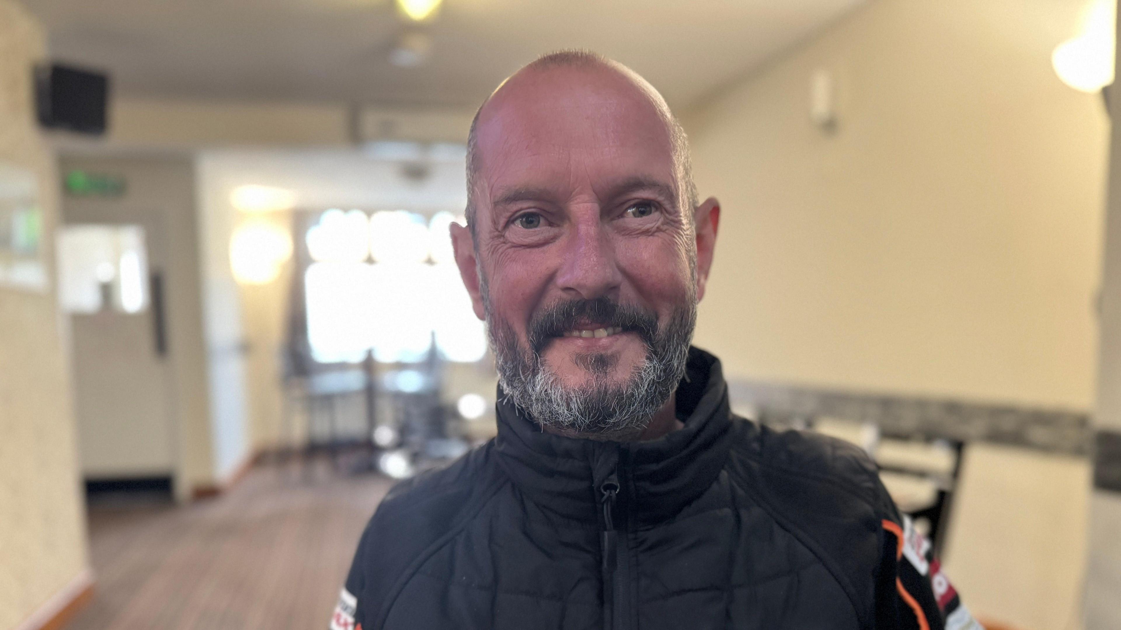 A man with a salt and pepper beard and very short hair. Wearing a black jacket zipped up to the top with logos on the sleeves. In a pub setting.