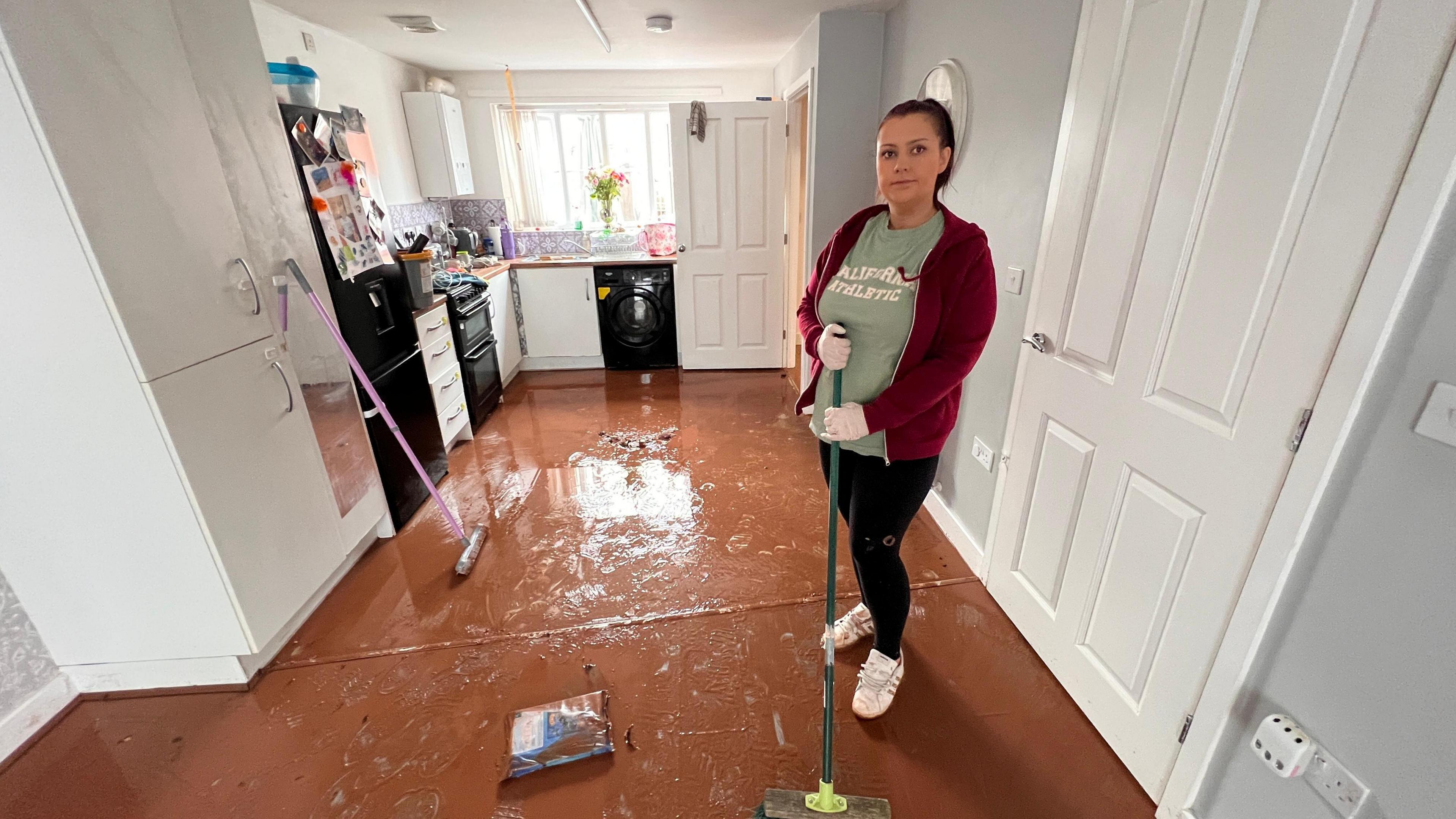 A woman holding a mop stands in a kitchen which is covered in brown sludge following flooding.