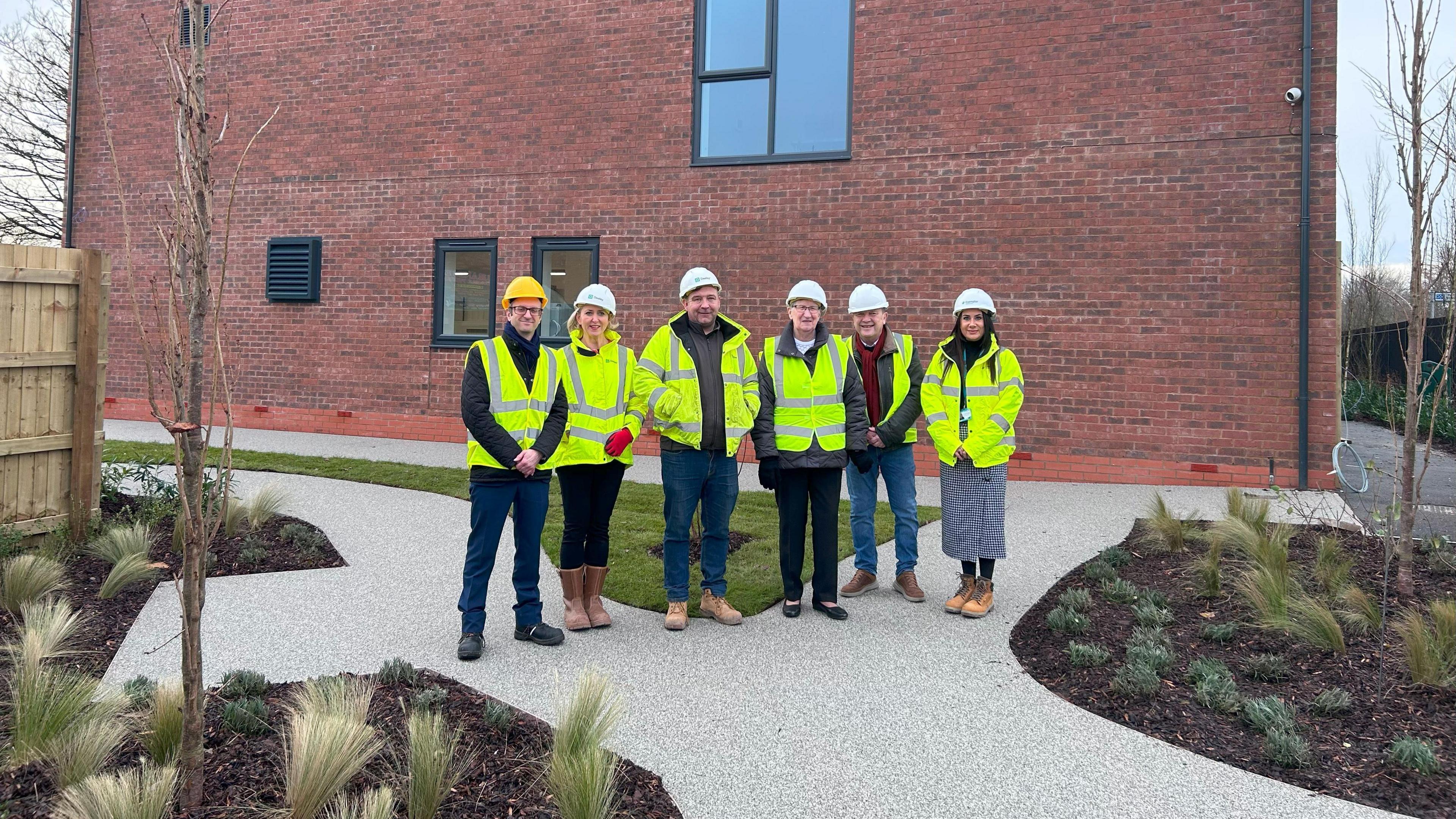 A group of six people are stood on a paved footpath, which is interspersed with several flowerbeds and a lawned area. The people are each wearing fluorescent yellow jackets as well as hard hats. Behind them is a large brick built building.