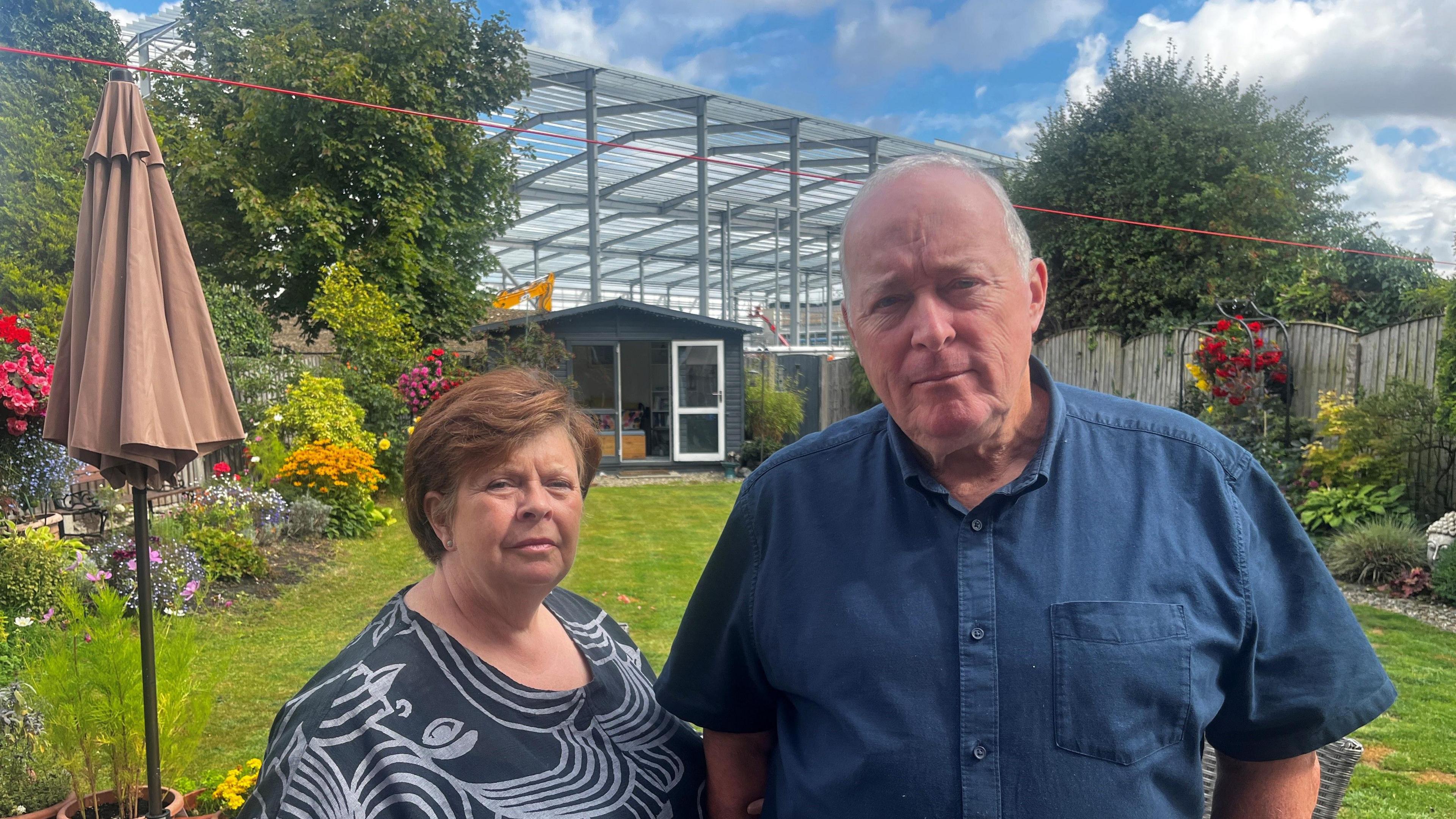An husband and wife stand in their grassy back garden. She is wearing a black and grey top and has short brown hair. He has short grey hair and is wearing an open-necked blue shirt. The metal foundations of a large warehouse loom over their back fence.