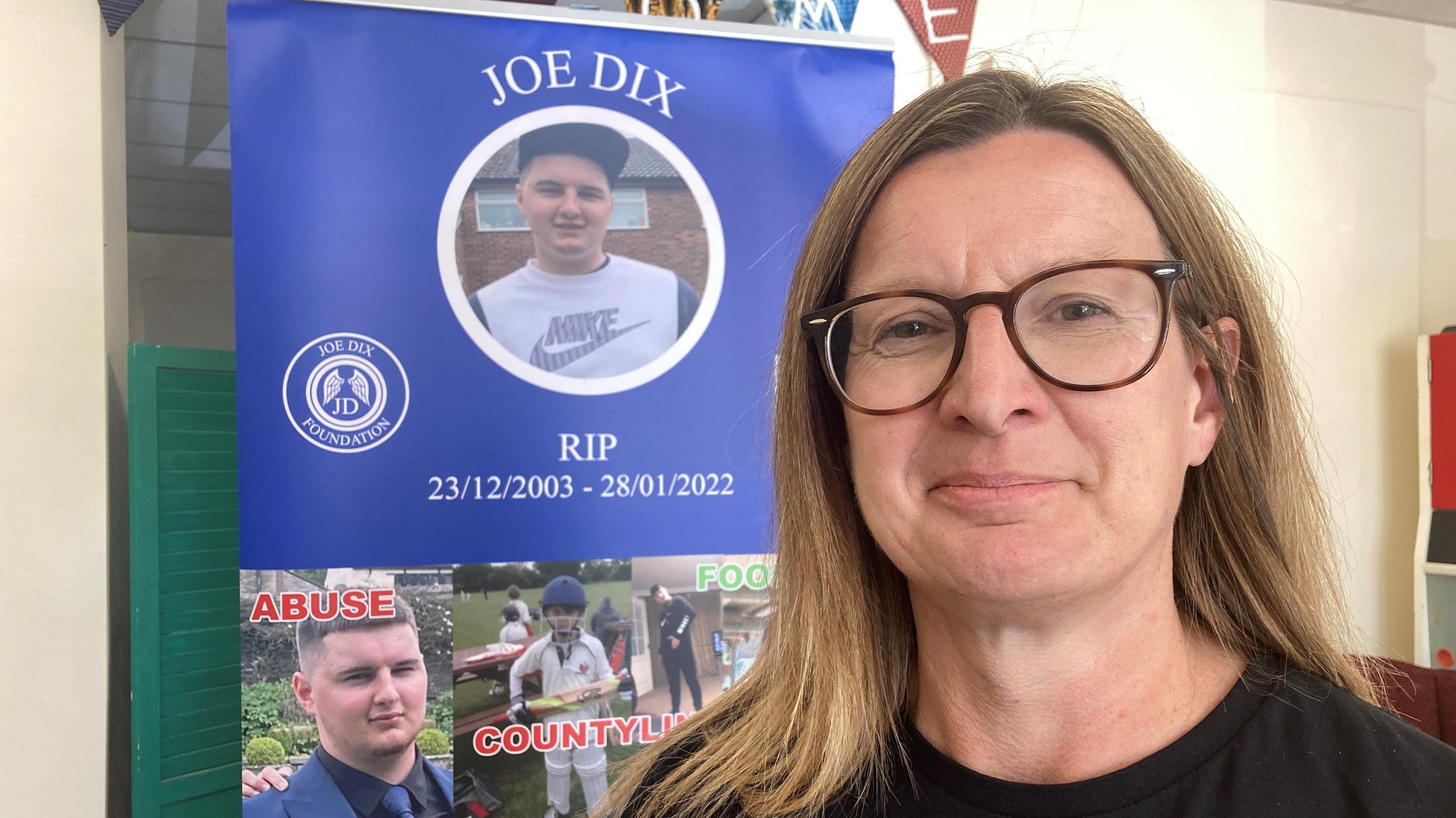 Emma Dix is on the right, looking into the camera and smiling. She is a brunette with shoulder-length hair, and she is wearing rounded glasses and a black t-shirt. To her left, there is a poster for the Joe Dix Foundation, which features images of Joe.