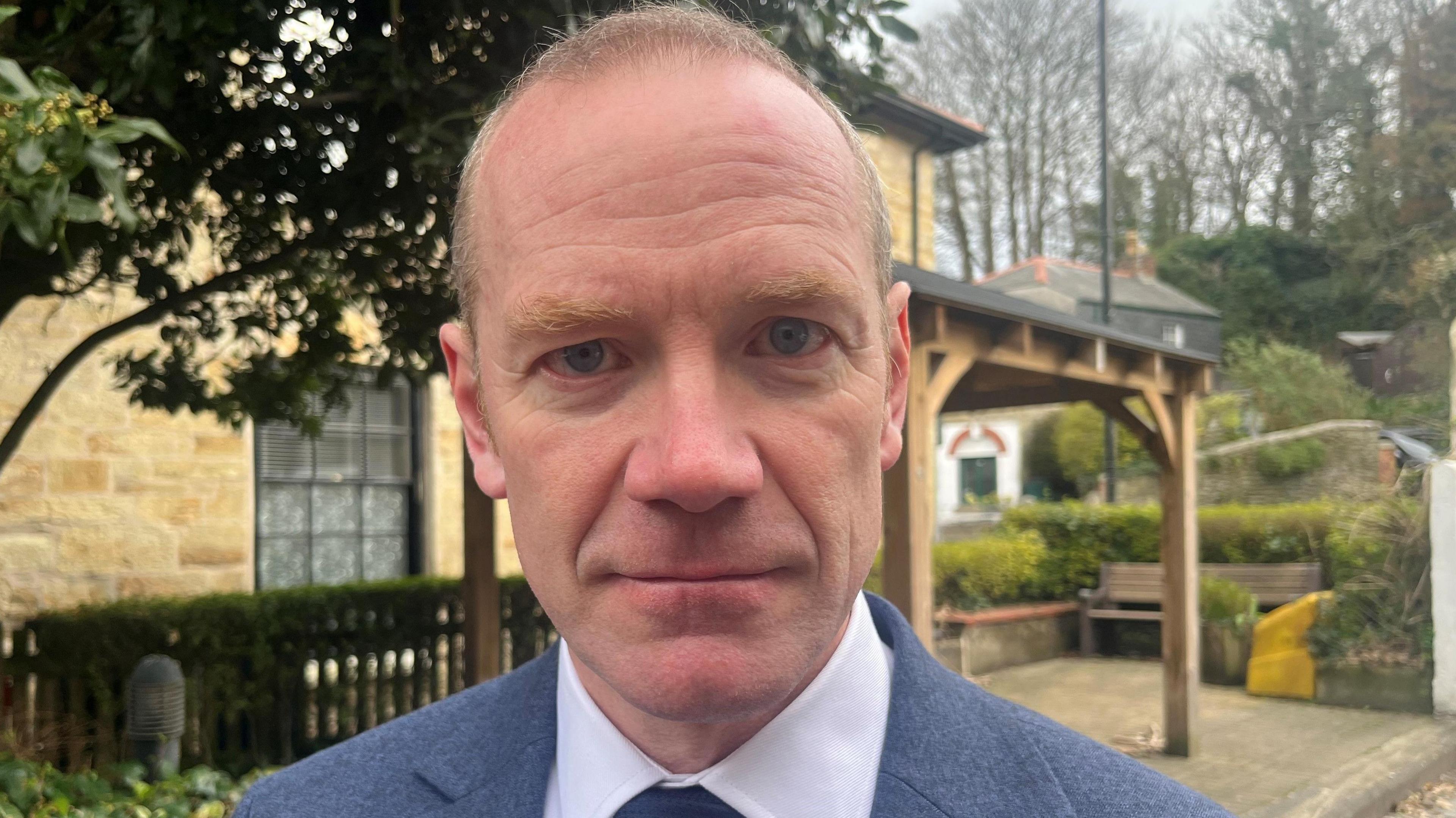 The head and shoulders of a man looking directly at the camera, he has thinning fair hair and blue eyes and is wearing a blue suit, white shirt and dark blue tie. The background is of a house and a tree.