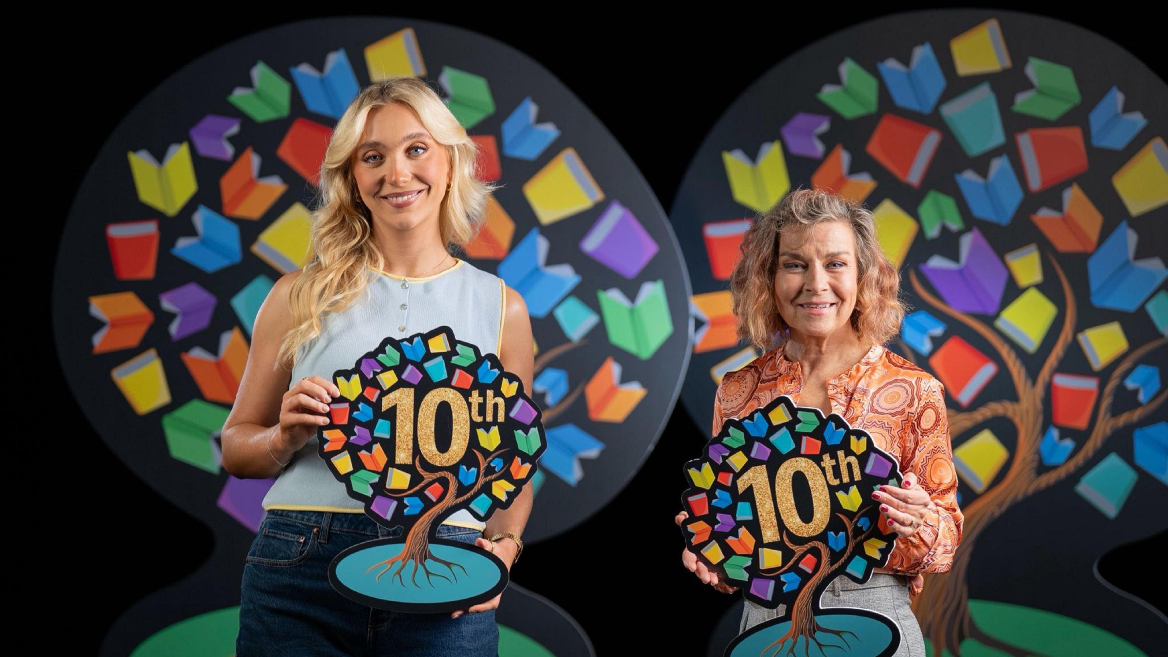 Ali and Cecilia are both holding Book Week symbols, which are in the shape of a tree with multi-coloured leaves and the word 10th in the centre. Ali has long blonde hair and is wearing a blue sleeveless top. Cecelia has shoulder length hair and is wearing an an orange patterned blouse.