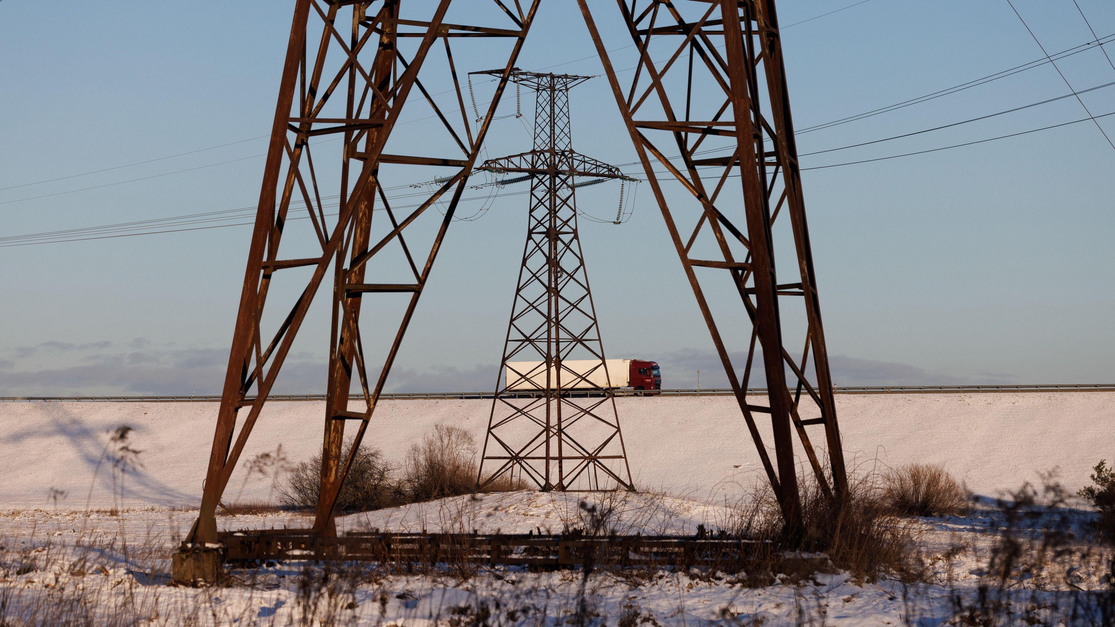 The bottom of one pylon in the foreground, with another pylon further away and a lorry on a road behind them both. There is snow on the ground and the sky is blue