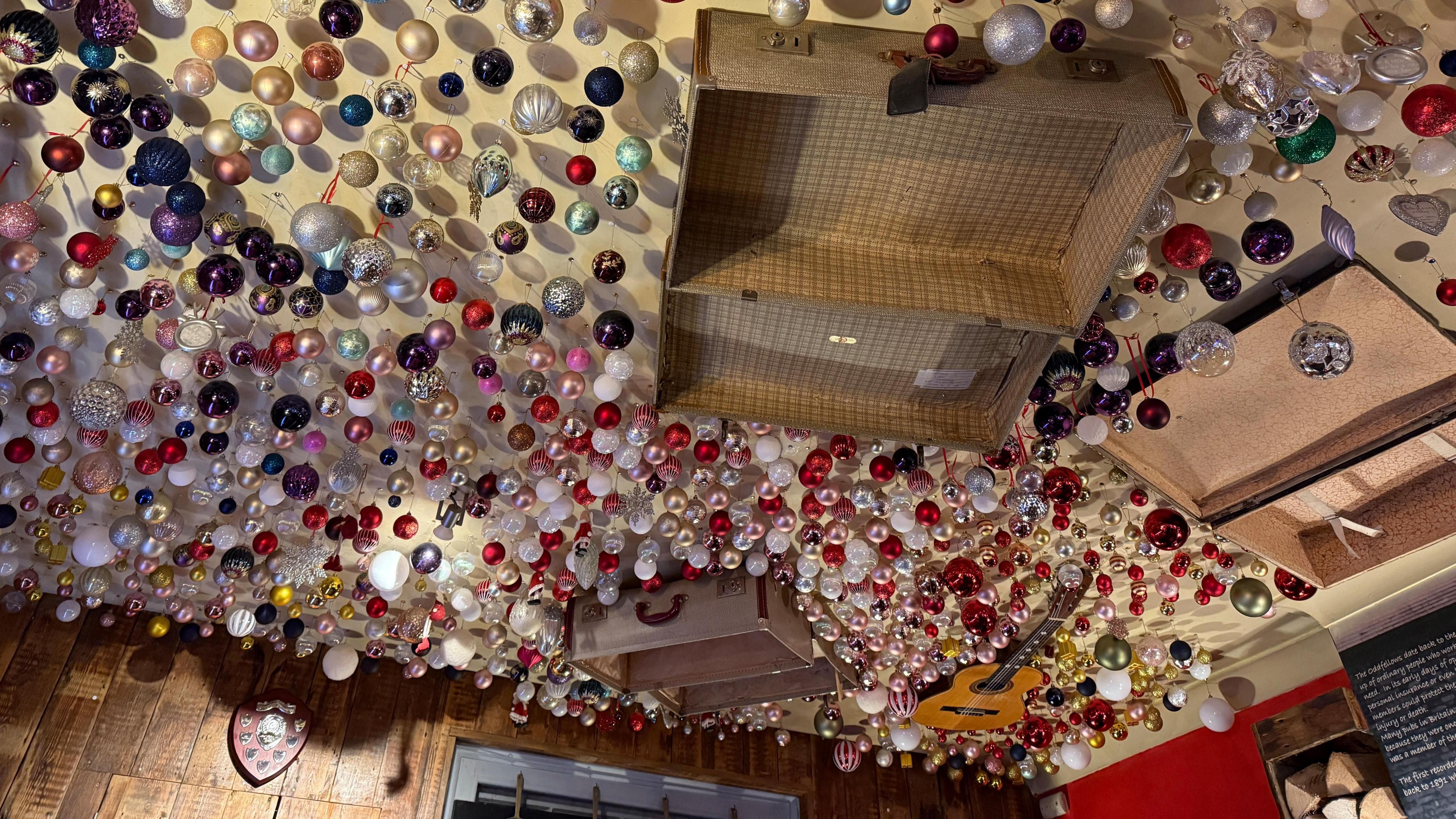 The ceiling of a pub decorated with hundreds of baubles. It also is decorated with old open suitcases and a guitar.