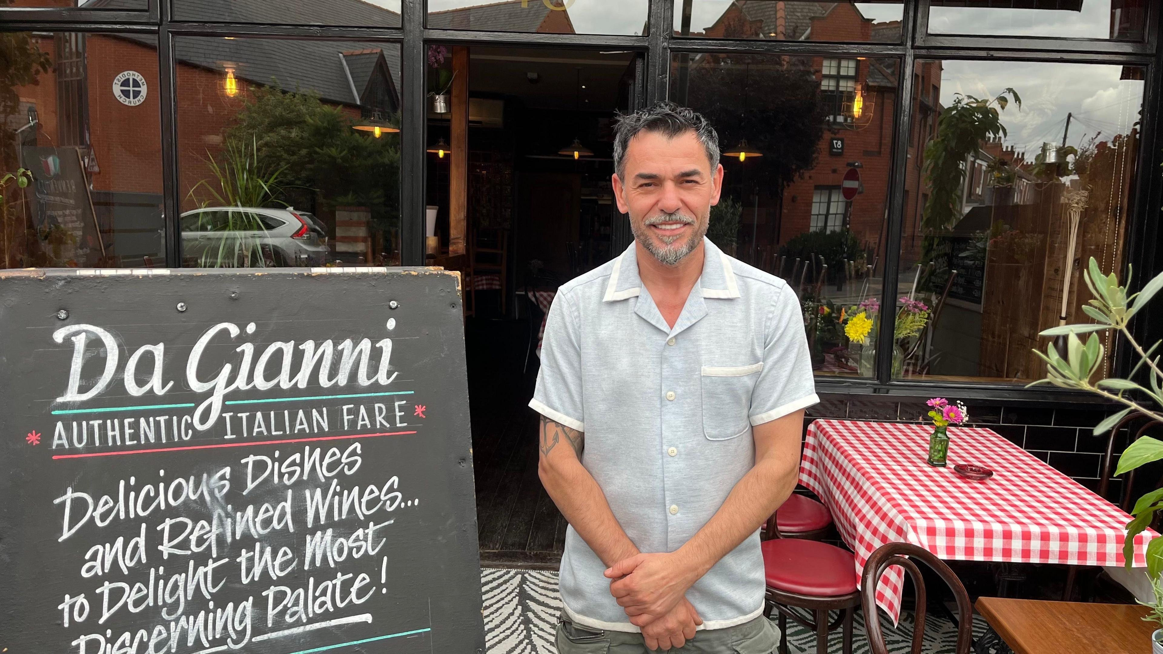 A man with a blue shirt, grey hair and goatee beard stands in front of a restaurant with large windows. A table has been covered with a red and white checked cloth and a large sign reads "Da Gianni - Authentic Italian Fare".