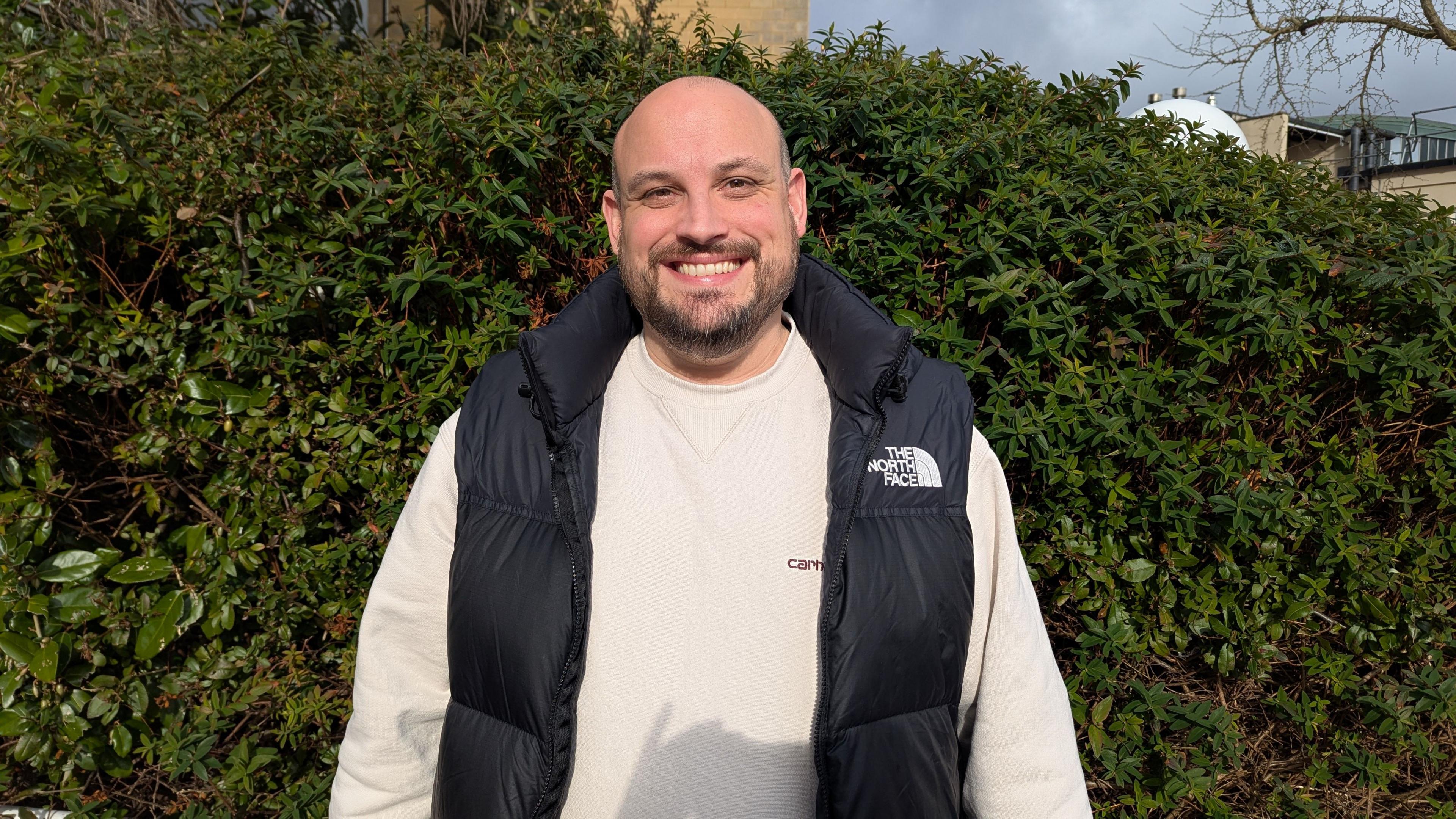 Pete Fealey wearing a white jumper and a black gilet standing smiling in front of a green hedge. 