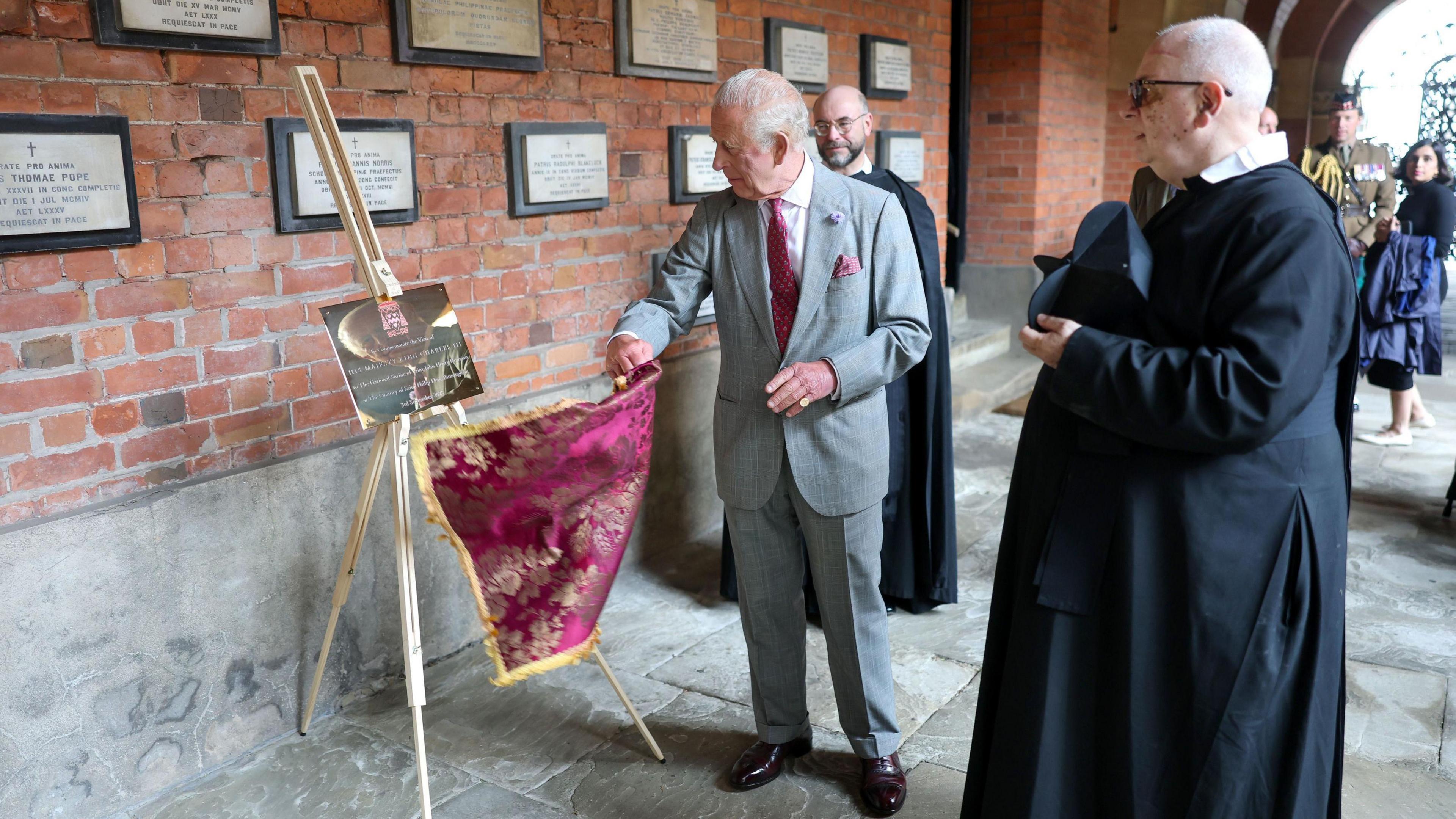 King Charles in grey suit and pink tie pulling an ornate pink cloth from a gold plaque on an easel. Other gold plaques line a red brick exposed internal wall. A Father in a black costume holding a black hat looks on, with other people visible behind the King.