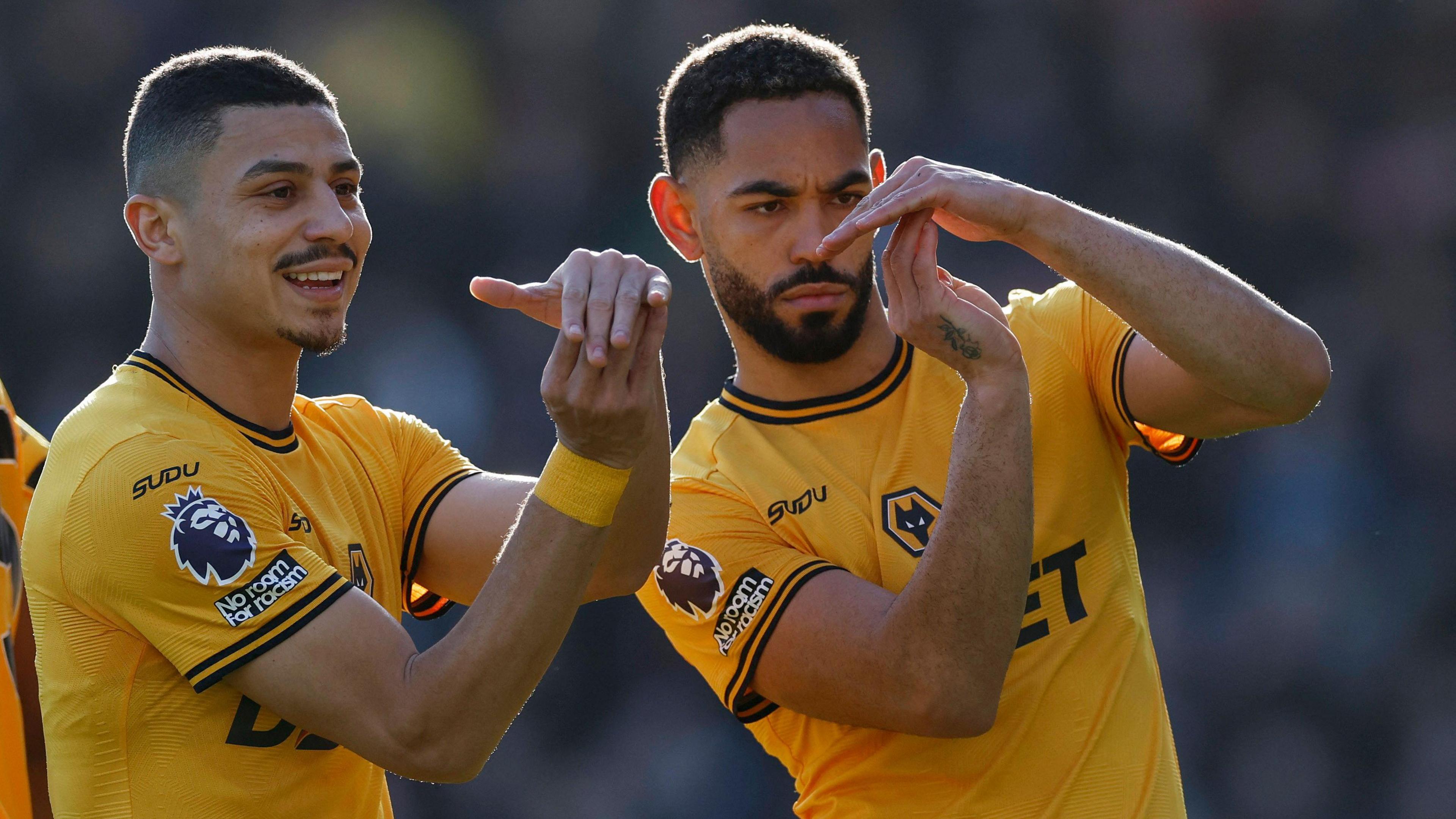 Matheus Cunha and another footballer to his right are wearing yellow Wolverhampton Wanderer shirts and gesturing the letter J during the game.