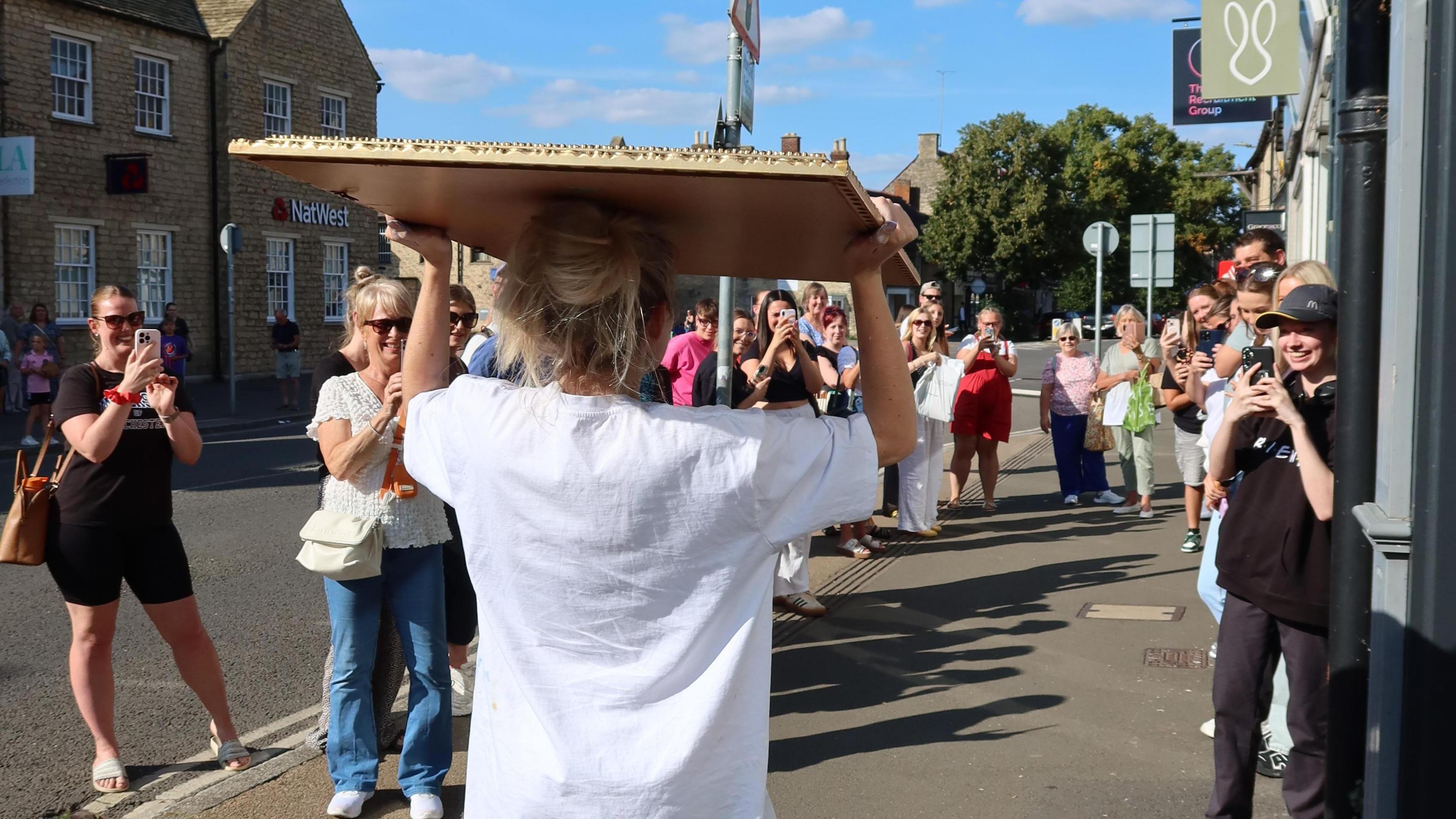 A blonde woman in a white t-shirt carrying a mirror on her head as she walks down the pavement with about 20 people watching and taking photos on their phones.