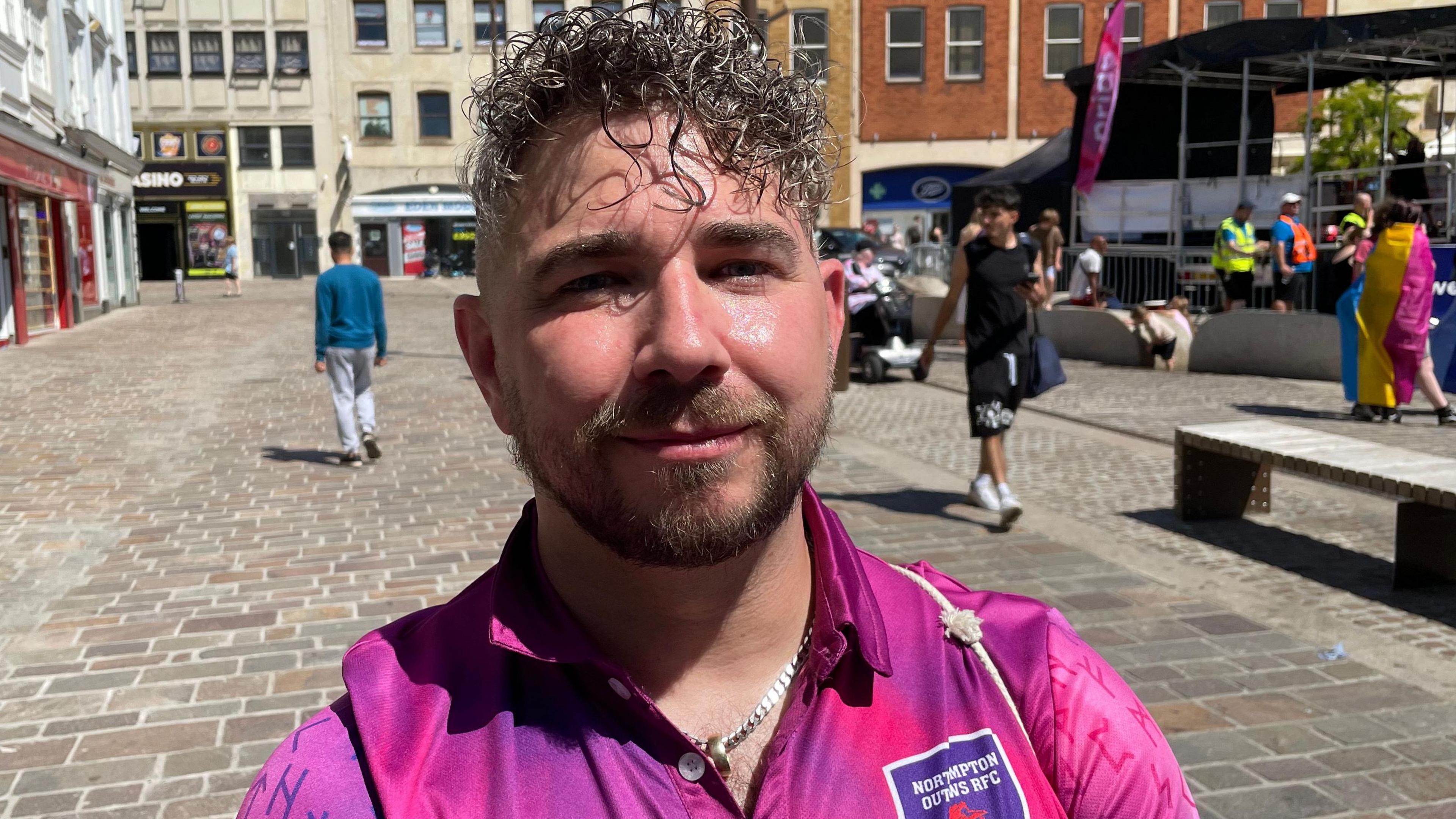Joey is standing in the sun while wearing a pink top with the logo for his rugby club. He is also wearing a chain around his neck. He has short curly hair. 