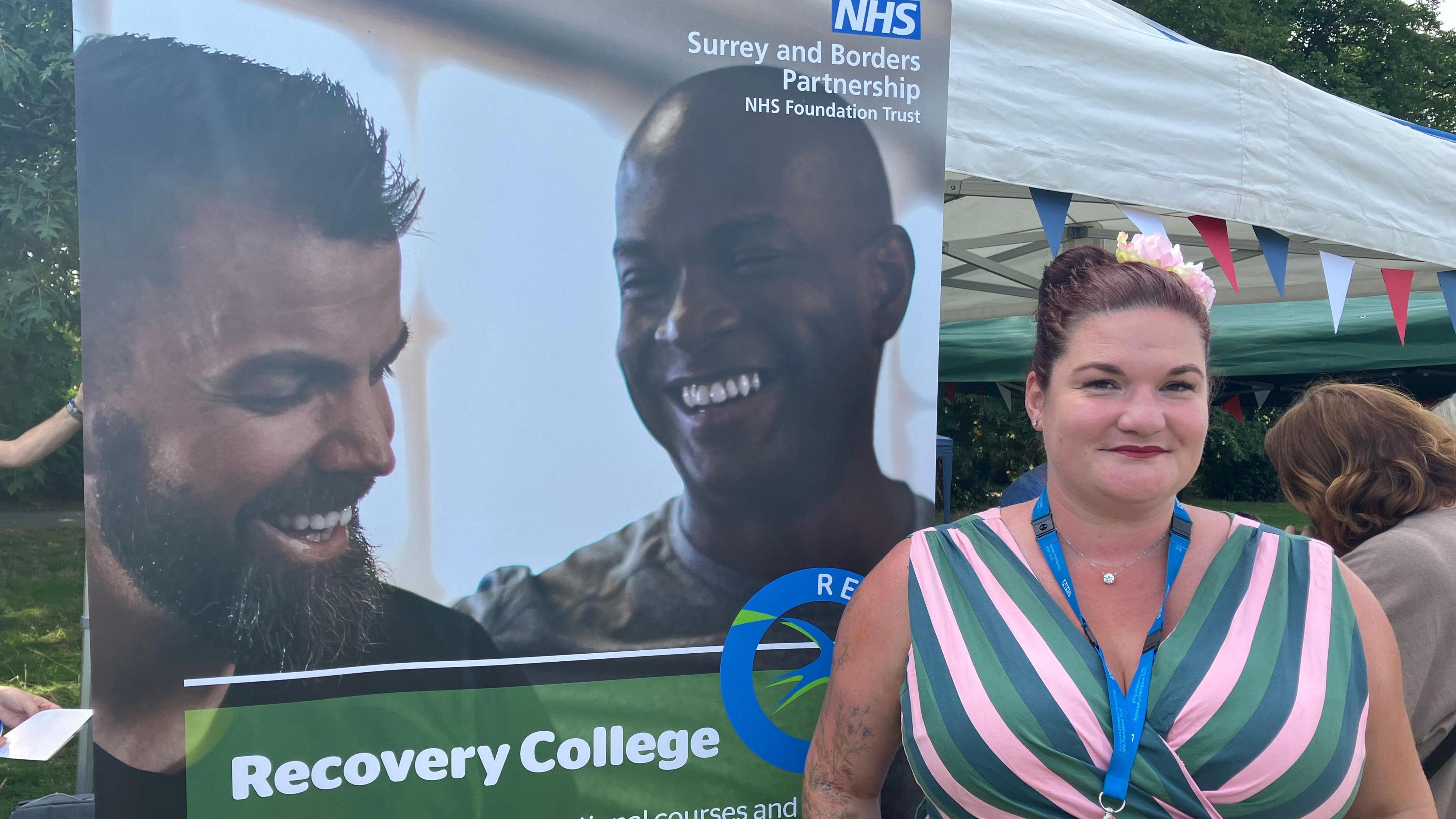 The Recovery College's service manager, Nina Clayton Broom, pictured in front of a poster advertising the service. Nina is wearing a lanyard and a green/pink dress. Two men are seen smiling on the poster.
