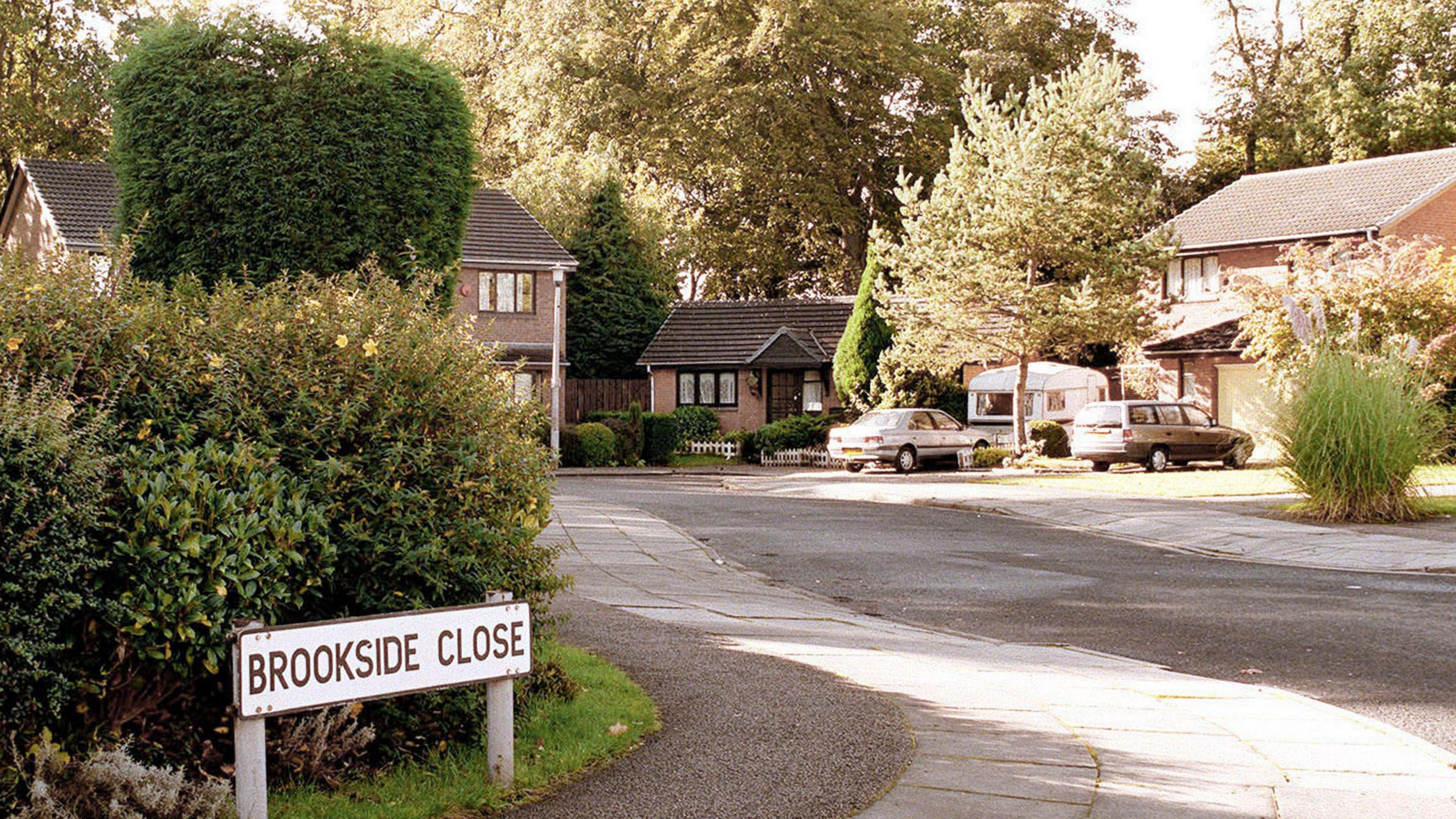 A picture of Brookside Close, captured in the 1990s. To the left is a street sign which reads: Brookside Close with bushes behind it. The road curves round to the left with a number of houses and one bungalow in view.