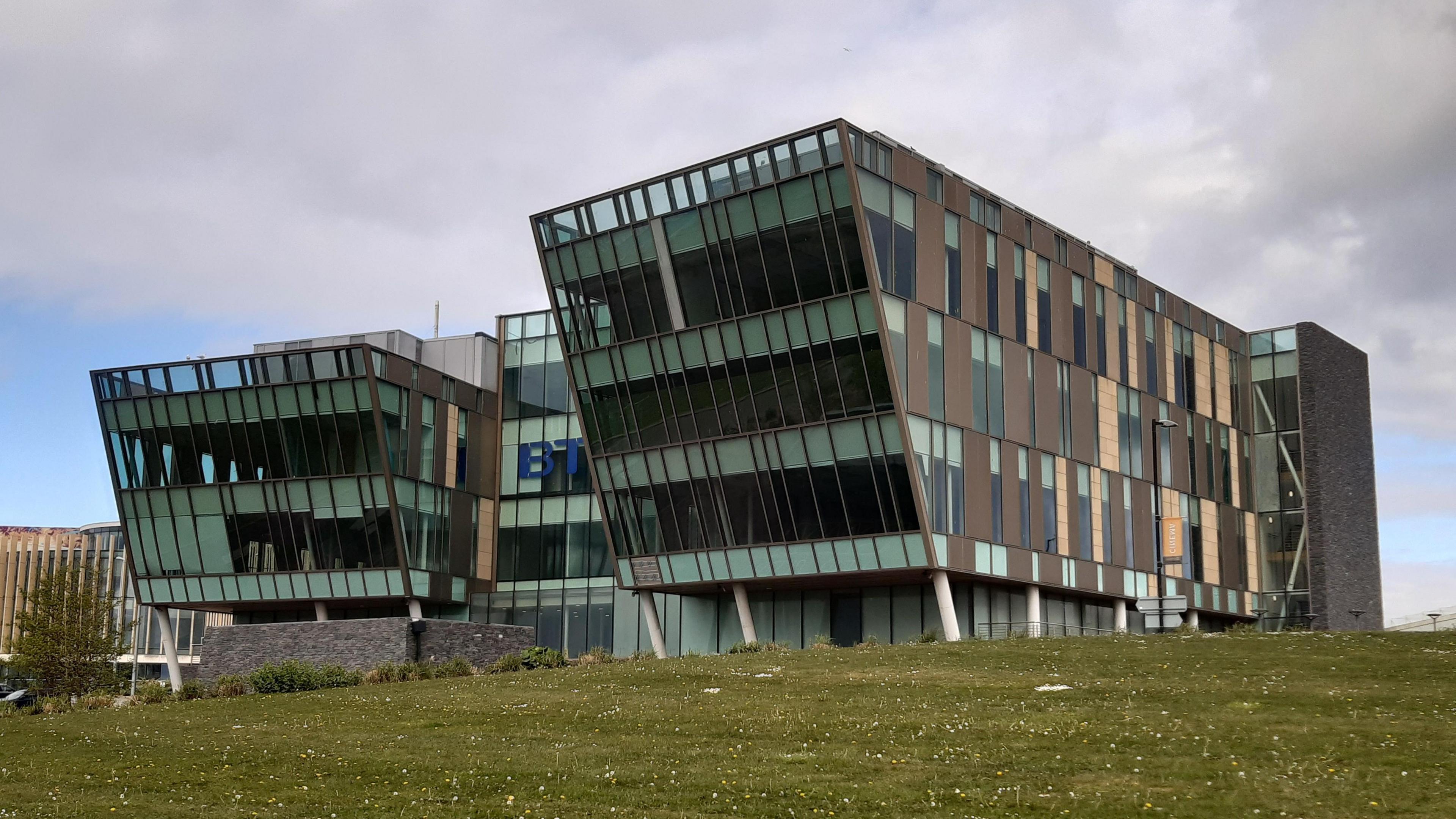 A large three-sectioned building, with brown panelling and glass frontage, with a blue BT sign in the middle. The fronts of the two end sections are sloped outwards so the top floor juts out further than the lower floors. There is a grass area in front of the building which leads down to the River Tyne, although that is out of shot. It is a cloudy but dry day.