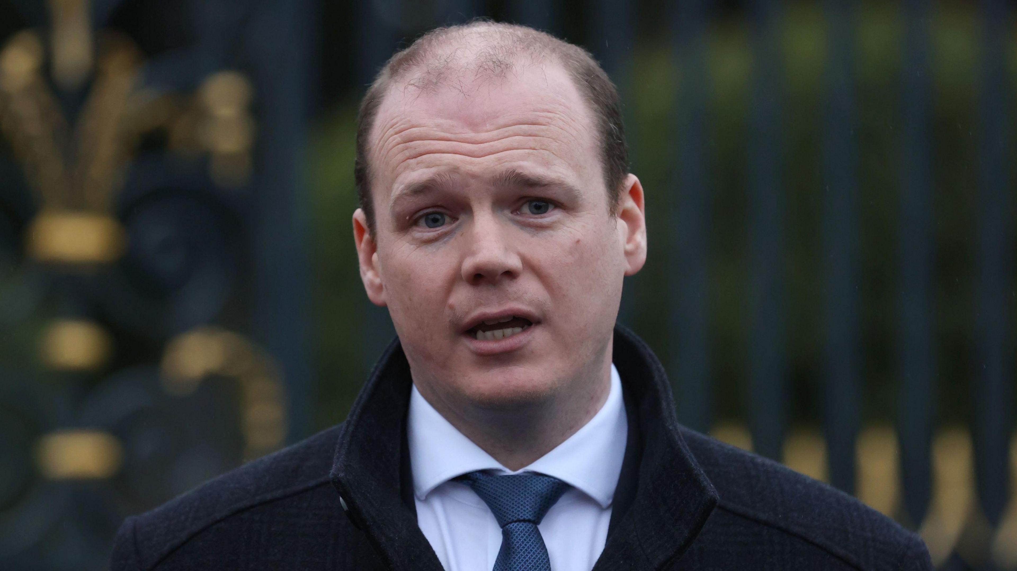 Gordon Lyons - a man is mid speech as he stands in front of a gold and black iron fence. He is wearing a black coat, with a white collared shirt and a navy tie.